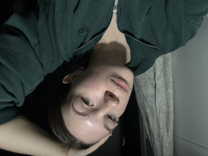 The image displays a single female individual in an indoor, dimly lit environment. The subject possesses light skin, greenish eyes, and light brown hair. She is wearing a dark green, possibly faded, full-zip sweatshirt featuring a silver zipper and a small, embroidered logo on the left chest. Her right hand is positioned on top of her head, with fingers visible. The individual's head is slightly tilted, and she is looking directly towards the camera. The background on the viewer's left consists of a light-colored door or wall and a hanging, light-toned, patterned fabric, possibly a towel or curtain. The background to the viewer's right is predominantly dark. Illumination appears to be artificial, casting highlights on the subject's forehead and skin, suggesting a light source from the front-left perspective. The specific location within the city of Vozdovac Urban Municipality, Serbia, cannot be ascertained from the visual evidence present in the image.