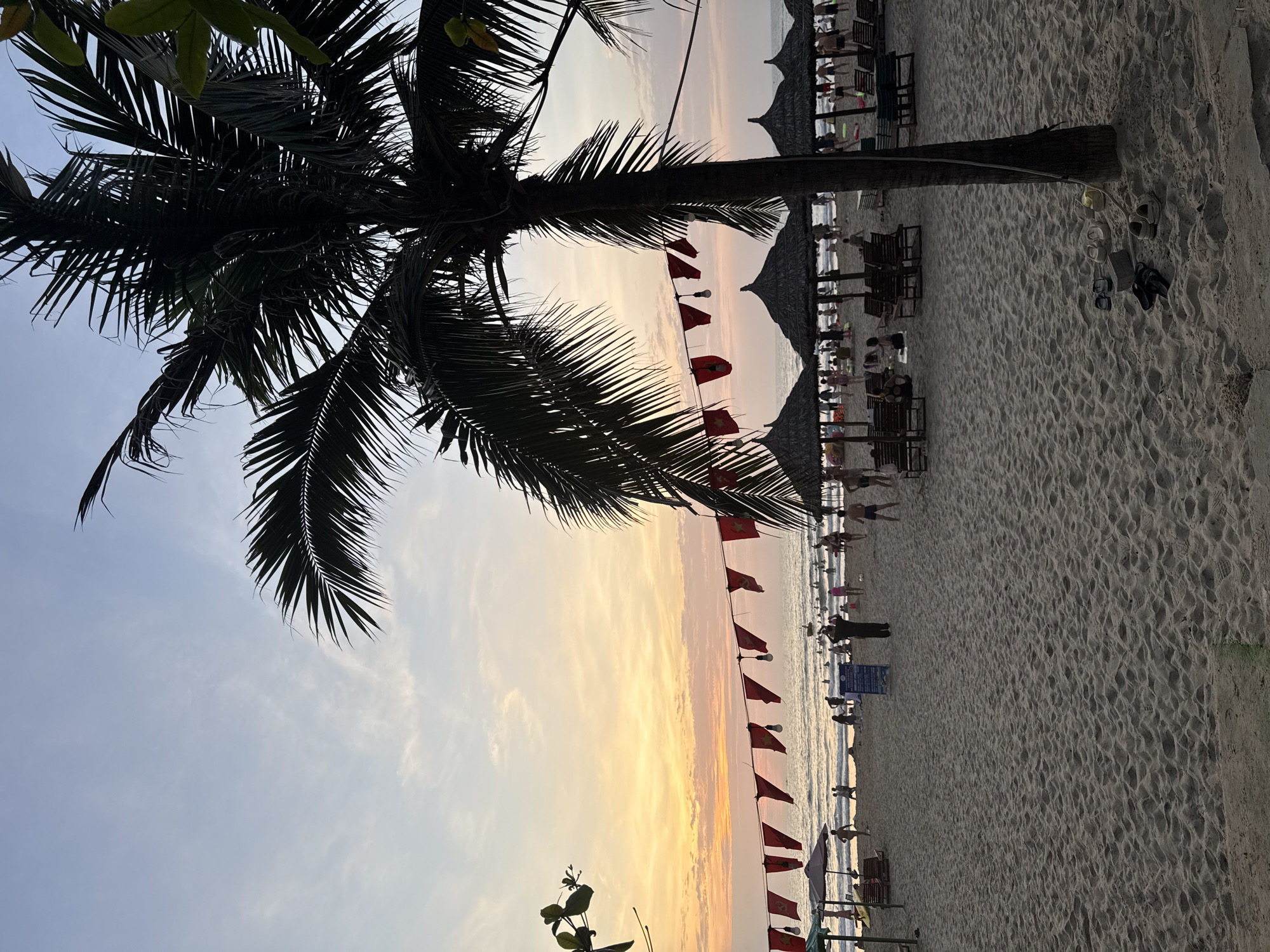 The image shows a beach scene in Đà Nẵng, Vietnam, at sunset. A palm tree is silhouetted in the foreground. A string of red flags hangs along the beach. In the background, people are relaxing under thatched umbrellas and on lounge chairs. There are also several pairs of shoes sitting on the sand. The sky is a mix of orange, yellow, and blue colors, indicating the sun is setting.