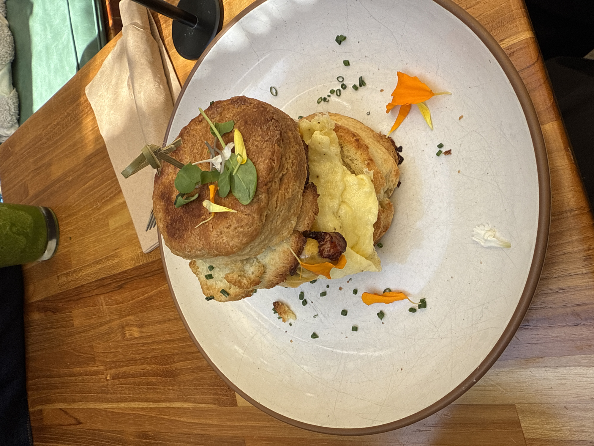 The image shows a plate of food on a wooden table. The food appears to be a breakfast sandwich, with two halves of a bun sandwiching a filling that includes what looks like scrambled eggs or melted cheese. The sandwich is held together with a small wooden skewer. There are small green sprigs, possibly chives, scattered around the plate along with small orange and white flower petals. A light-colored cloth napkin is visible to the left of the plate, with what appears to be a glass of green juice also on the left side of the frame. The image does not provide enough information to determine the location in Los Angeles, United States of America, or to determine any people or interactions.