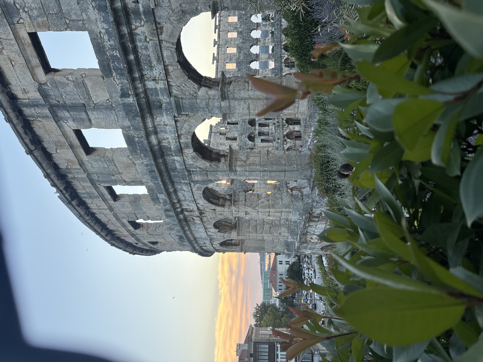 An exterior shot of the Pula Arena at dusk. The amphitheater, constructed of stone, curves from the right side of the frame towards the left. Its upper levels feature rectangular openings, while the lower levels are characterized by a series of arched walkways. In the foreground, lush green foliage, some with reddish tinges, is out of focus, creating a natural frame for the ancient structure. Beyond the amphitheater, a cityscape is visible with buildings and a parking lot filled with cars. The sky above displays a soft gradient of pale blue transitioning to warm orange and pink hues near the horizon, indicating the setting sun. A few birds are captured in flight against the sky. A person is partially visible in the middle ground, among the vegetation and behind the amphitheater's arches.