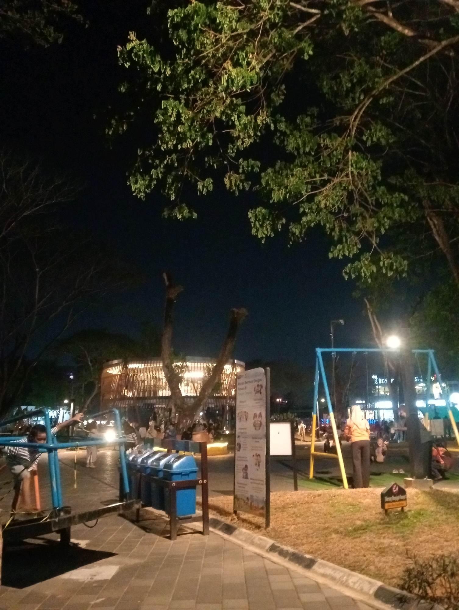 A nighttime scene at a playground and outdoor seating area, likely in Samarinda, Indonesia. The foreground features a paved walkway with a black and white curb, leading to a playground with blue and yellow swing sets. Trash receptacles are visible near the playground equipment. A young person in a striped shirt is interacting with a piece of playground equipment. In the mid-ground, a multi-story, modern structure with open wooden latticework and illuminated interior is visible, suggesting a restaurant or community space. Several individuals are scattered throughout the area, some sitting and others standing. Trees with green foliage are prominent in the upper portions of the image, and the night sky is dark.