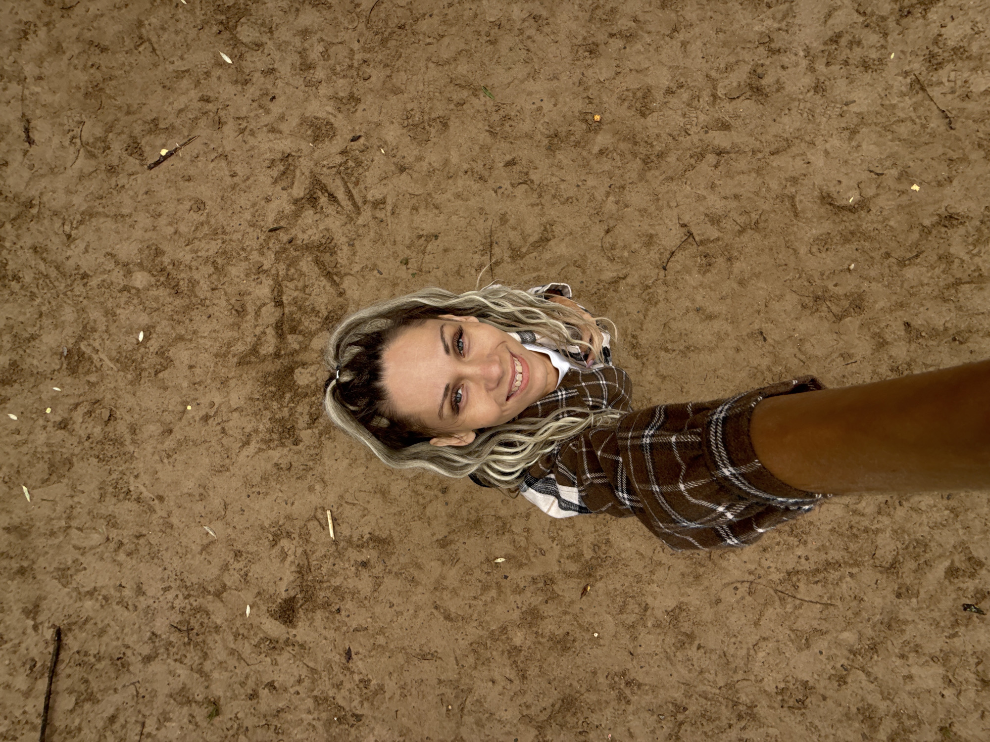 A first-person perspective, taken from a low angle looking up, shows a woman smiling at the camera. She has blonde, wavy hair pulled into a high ponytail with dark streaks. She is wearing a brown and white plaid collared shirt over a white t-shirt. The background is a dirt ground with scattered twigs and leaves. The image is set in an outdoor environment.