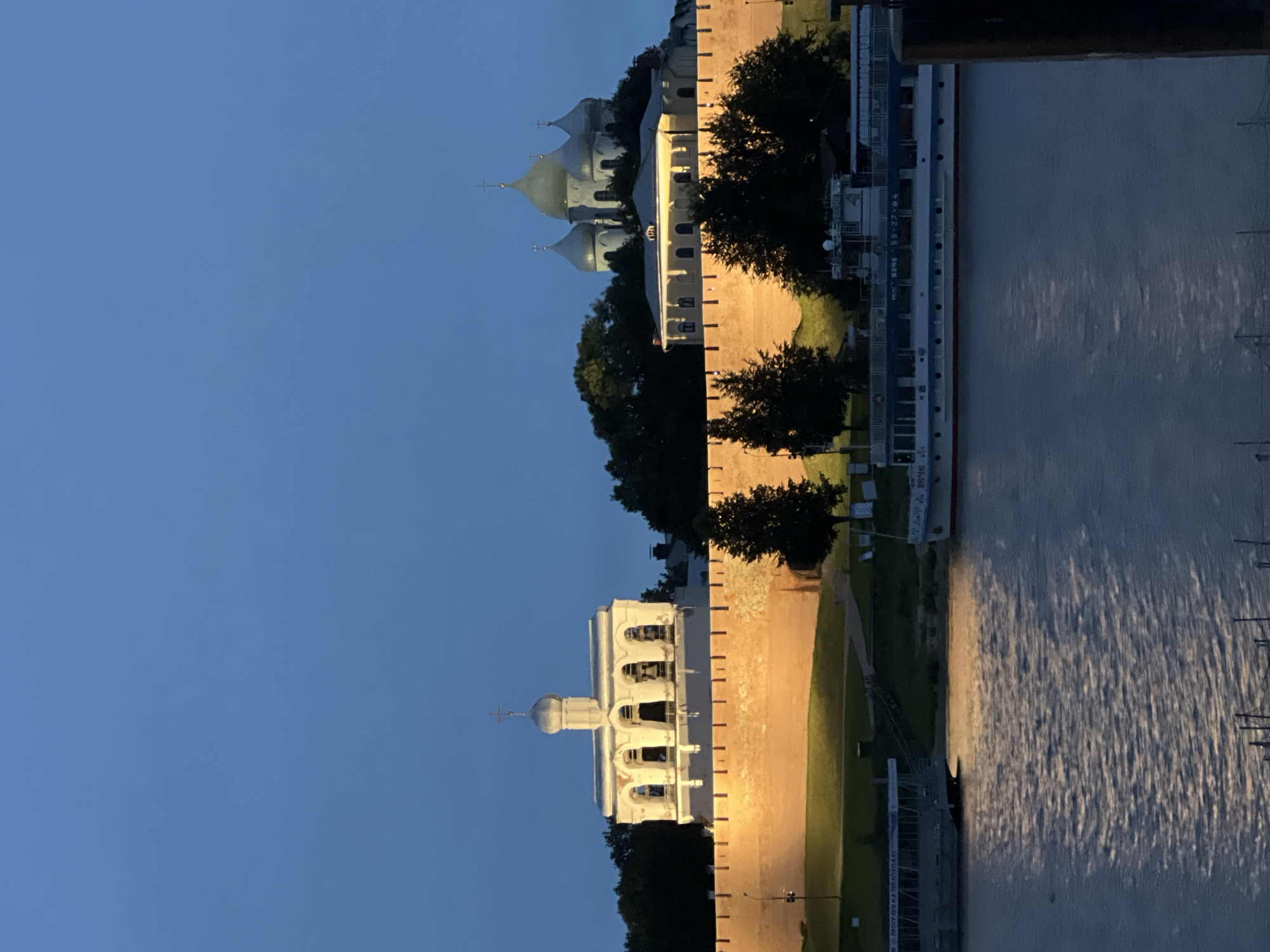 The image depicts a twilight view of the Veliky Novgorod Kremlin walls, illuminated by artificial lights from the riverbank. The walls, made of reddish-brown brick, stretch across the midground, topped with crenellations. To the left, a white bell tower with arched openings and a dome topped with a cross stands prominently, its bells visible within. To the right, behind the walls and trees, a church with multiple silver domes and crosses is silhouetted against the darkening sky. In the foreground, a wide river reflects the dim light, with several boats moored along the opposite bank. One larger, multi-decked passenger boat with white and blue detailing is clearly visible. The sky above is a deep, clear blue, indicating the end of daylight. The overall scene conveys a sense of historical grandeur and tranquility.