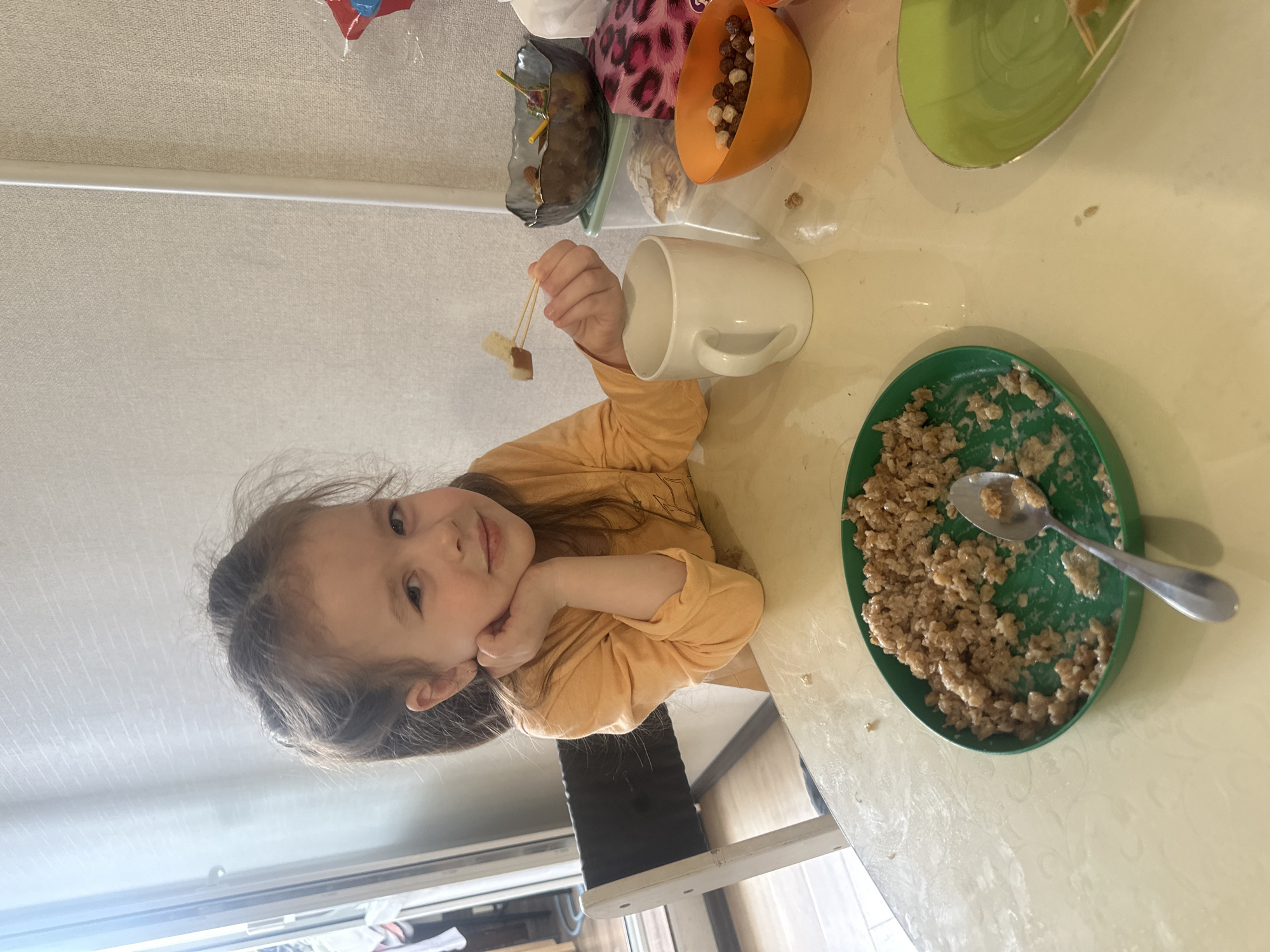 A young girl with dark hair, wearing a light orange long-sleeved shirt, is seated at a table, likely in a home in Tomilino, Russia. She looks directly at the camera with a slight smile, resting her chin on her hand, appearing content.
The scene depicts a mealtime, possibly breakfast. On the light-colored table in front of her, the girl holds a small skewer with two pieces of food, one light and one dark, in her right hand. A white ceramic mug sits nearby. Further back, an orange bowl contains a mix of chocolate and plain cereal puffs. Adjacent to this, a dark patterned bowl also holds small food items on skewers. To the right, a green oval plate is filled with what appears to be oatmeal or porridge, with a silver spoon resting in it. Another, mostly empty, light green plate is also visible. The background shows a light-colored wall or curtain, and a window or glass door provides natural light to the scene. The image is oriented with the top of the scene to the left and the bottom to the right. No visible text is present.