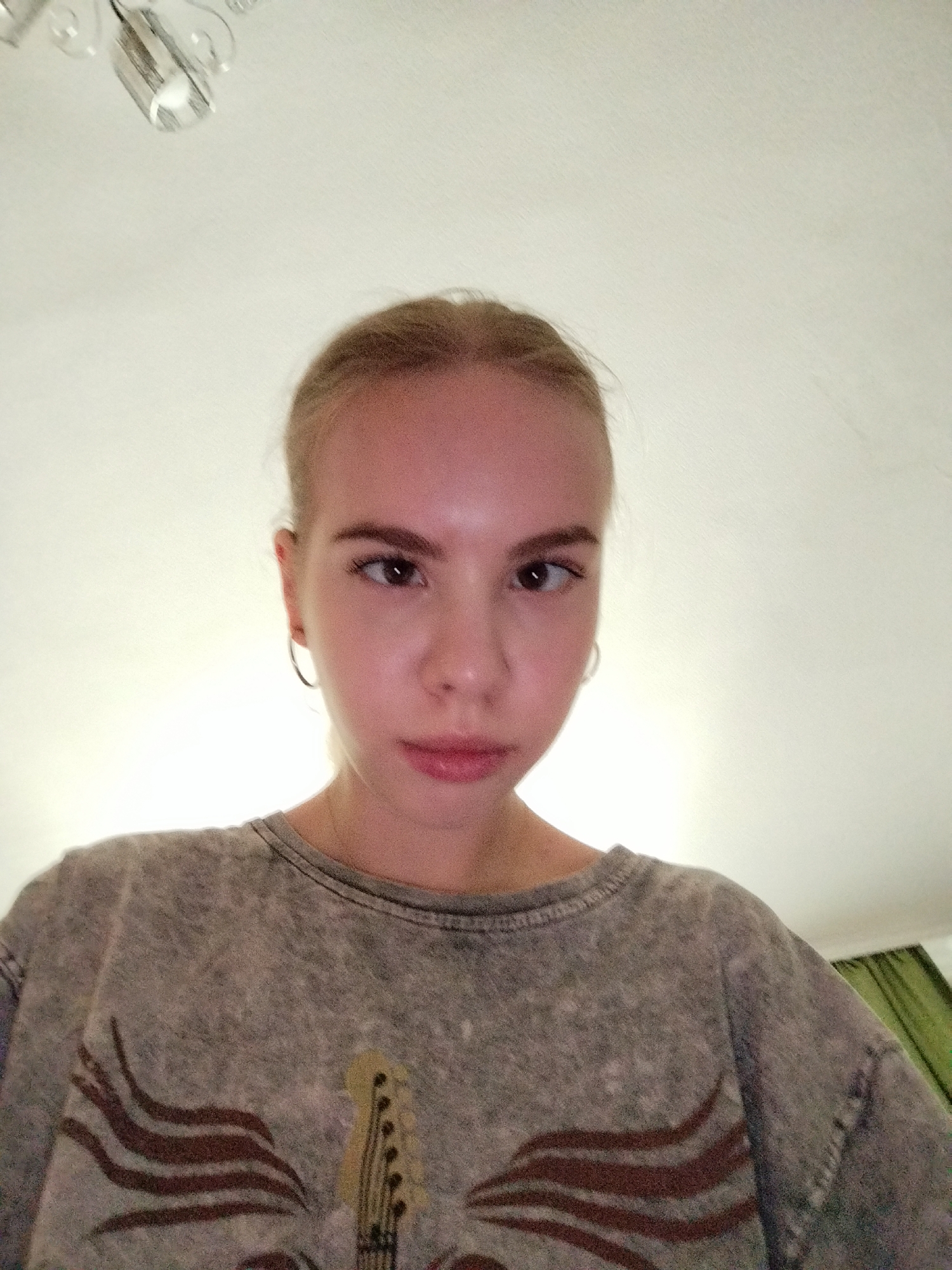 A young woman with blonde hair pulled back and a grey t-shirt with a graphic print of a guitar and flames is looking directly at the camera. She is wearing hoop earrings. The background is a plain, light-colored wall and ceiling. A light fixture is visible in the upper left corner of the image. The lighting appears to be artificial, suggesting the photo was taken indoors. There is no visible text in the image.