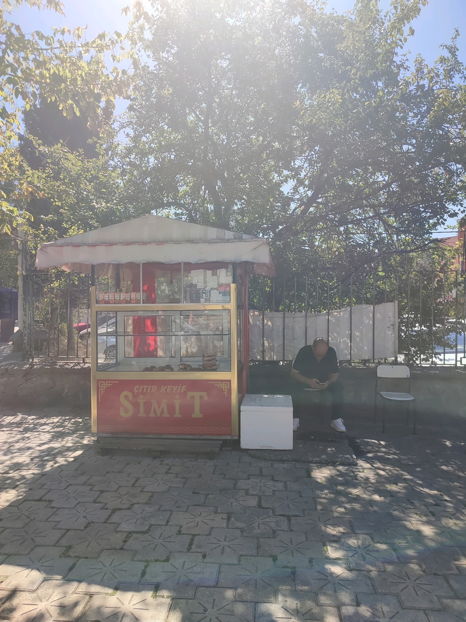 A man is sitting on the ground outside a "Simit" (Turkish bagel) stand in Kadıköy, Turkey, looking at his phone. The stand is yellow and red, with "ÇİTİR KEYİF SİMİT" written on the front. There are simit pastries displayed inside the glass case. The man is wearing a black t-shirt and shorts and is sitting next to a white cooler box. A white plastic chair is to his right. The scene is illuminated by bright sunlight, casting long shadows on the patterned stone pavement. Trees with green leaves are visible in the background, along with a metal fence.