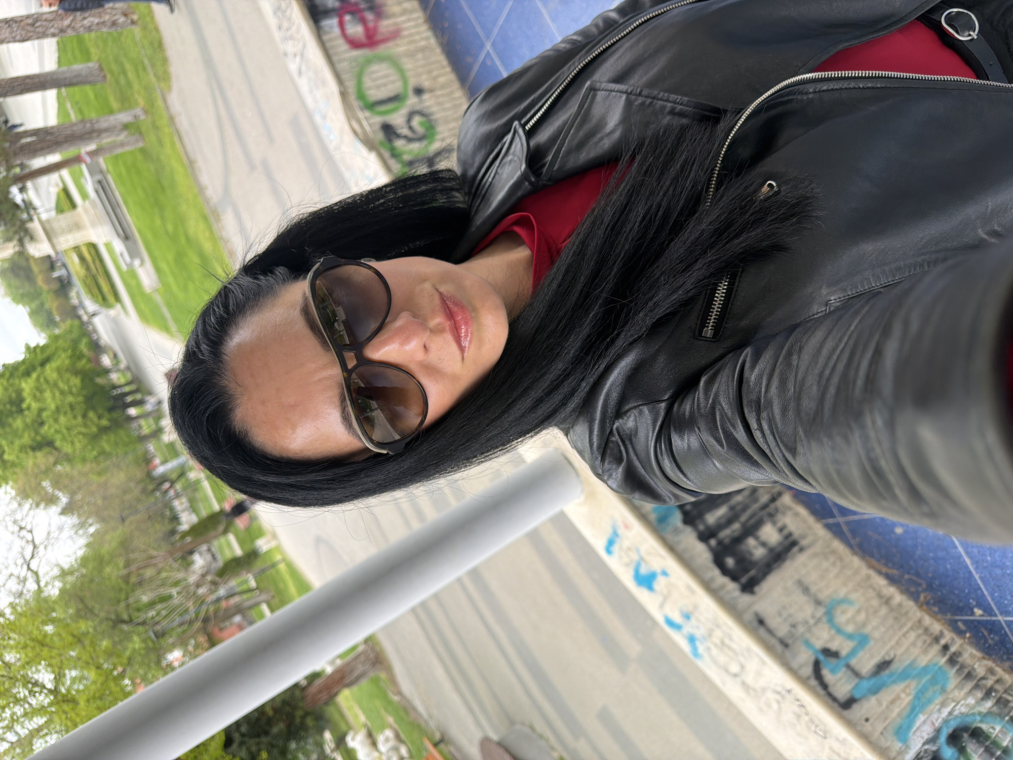 A woman with long black hair, wearing sunglasses, a red top, and a black leather jacket, is positioned outdoors. She appears to be standing near a low wall or barrier with graffiti on its surface. In the background, there is a park-like area with trees and a walkway visible, suggesting an urban or suburban setting. The photograph seems to have been taken in Pleven, Bulgaria.