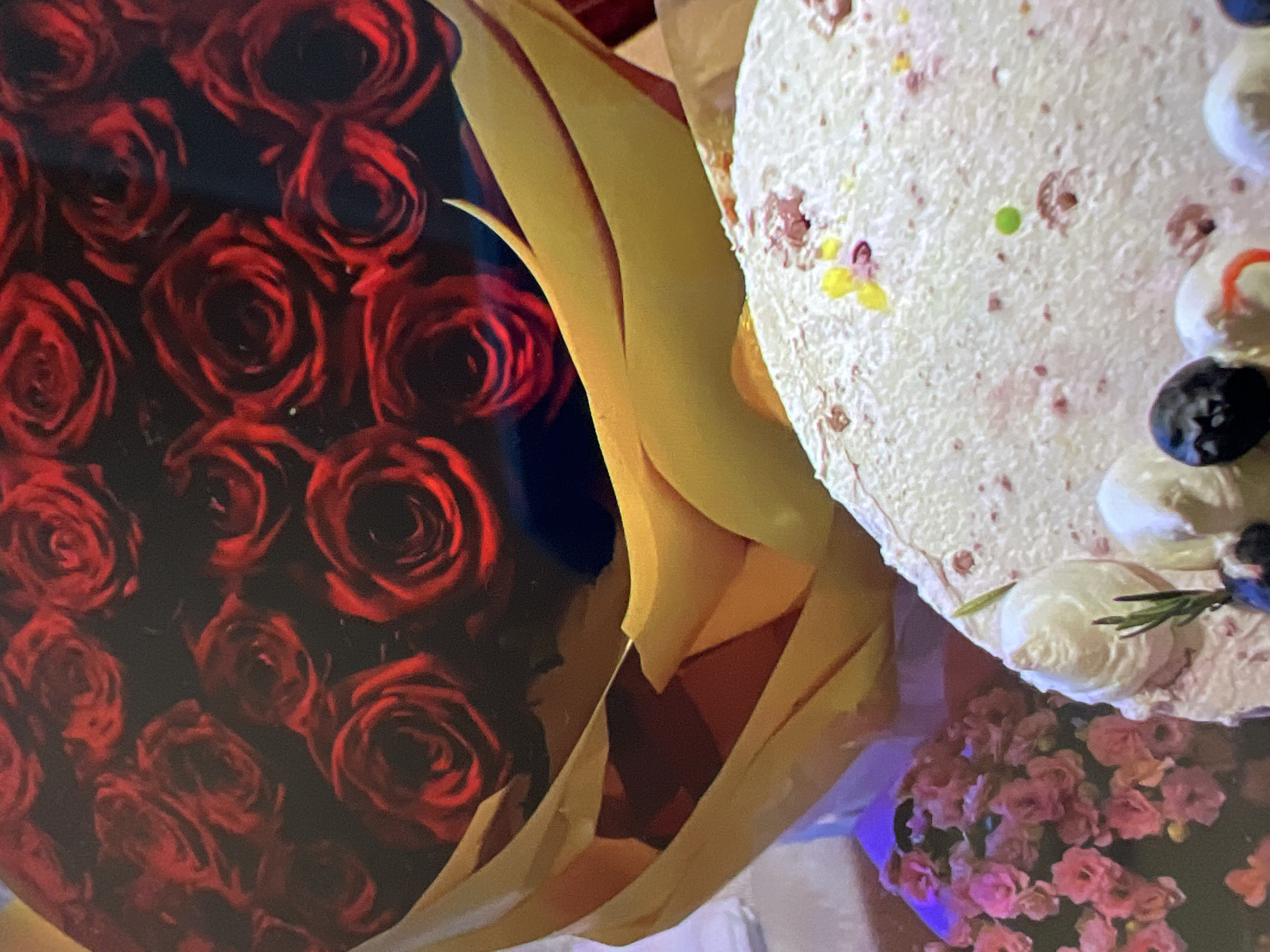 The image shows a close-up of a bouquet of red roses wrapped in yellow paper, and a portion of a pink cake decorated with white frosting and fruit. The bouquet of roses is in the upper half of the image, with the yellow paper wrapping forming a large, curved shape. The cake is in the lower right portion of the image, with its textured surface and various toppings visible. To the left of the cake, a small cluster of pink flowers is partially visible. The overall scene appears to be set indoors, possibly at a celebration or event. There are no people or discernible city elements present in the image.