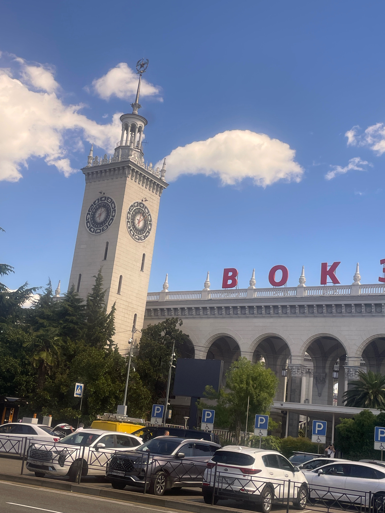 The image shows a sunny day in the city of Sochi, Russia, with a clear blue sky and fluffy white clouds. In the foreground, several cars are parked along a street, including white SUVs and a dark-colored SUV. Behind the cars, a building with a grand facade featuring arched windows and decorative columns is visible. Large red letters spelling "ВОКЗАЛ" (meaning "station" in Russian) are displayed on the top of the building. To the left of the building stands a tall, white clock tower with a spire and a star on top. The clock faces on the tower are circular with intricate designs. The area appears to be a public space, likely a train station, given the signage and the presence of multiple vehicles. Green trees and palm trees are scattered throughout the scene, indicating a temperate climate. A pedestrian crossing sign is visible on a pole.