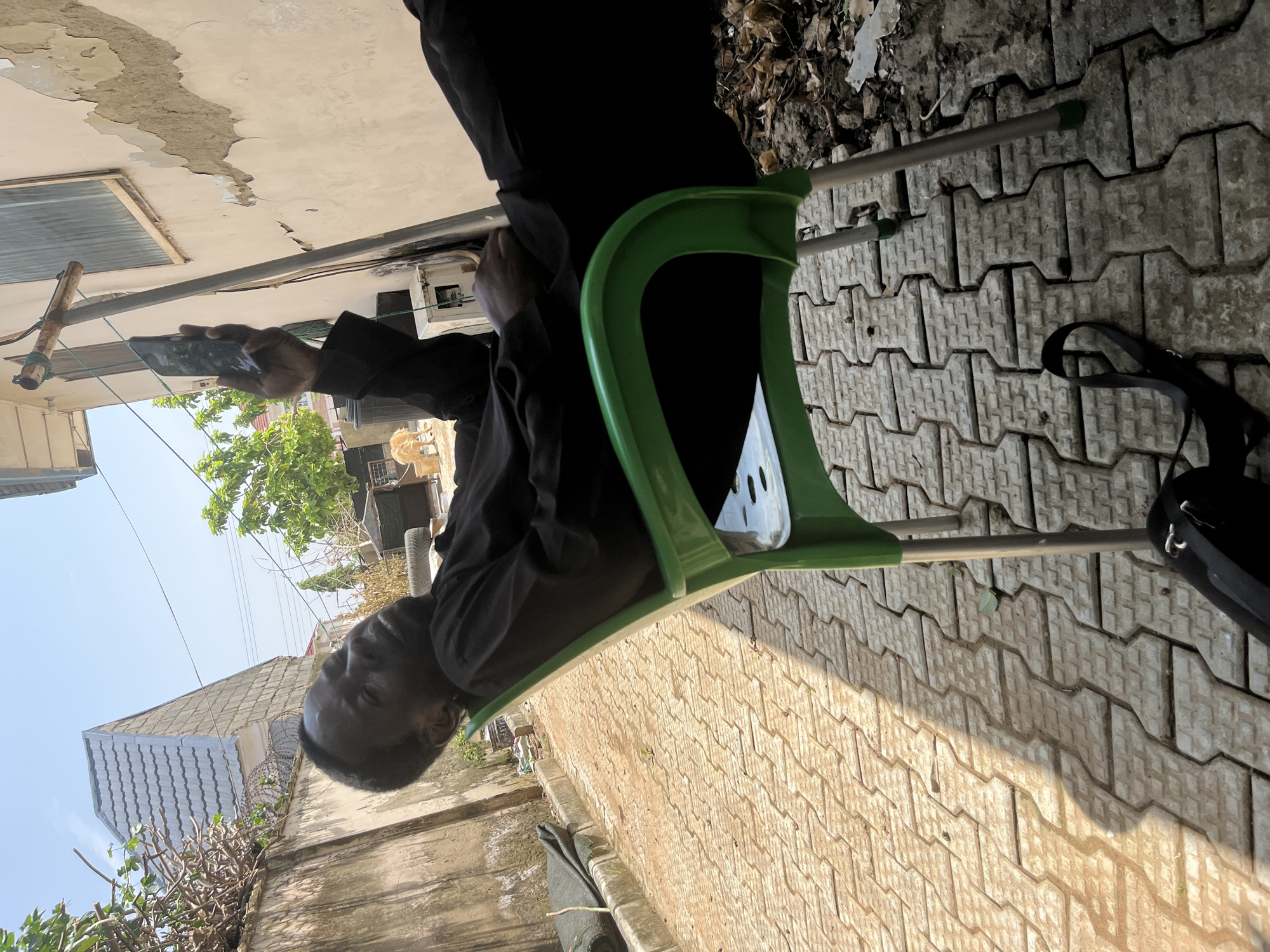 In Abuja, Nigeria, a person is seen reclined in a green plastic chair on a patterned stone patio. The individual, dressed in dark clothing, is holding a smartphone with their right hand, appearing to be looking at or using the device. The background reveals a slightly weathered wall with peeling paint, a portion of a building with a tiled roof, and some foliage. A black bag with straps lies on the patio near the chair. The scene suggests an outdoor, possibly residential, setting under daylight.