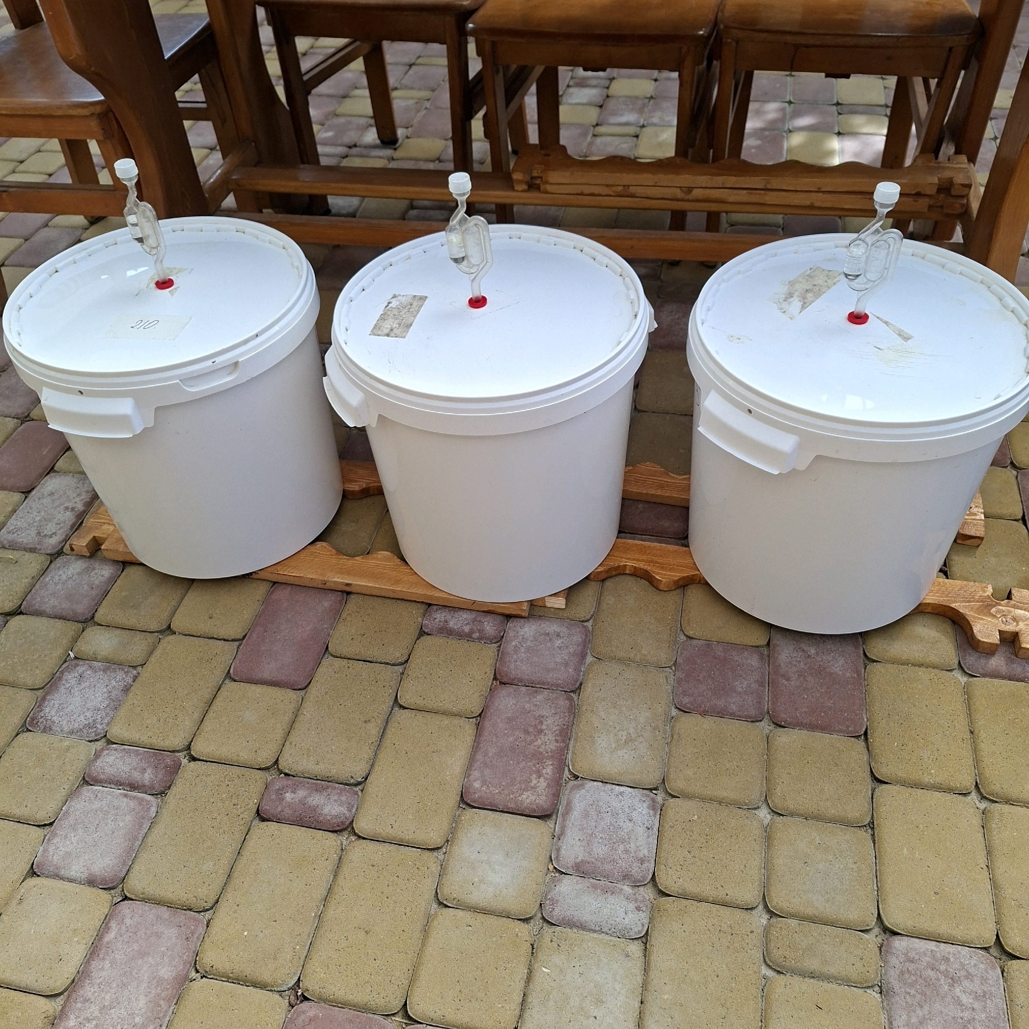 Three white plastic buckets with red-capped airlocks are placed on a paved patio area. The buckets are on wooden stands. In the background, wooden chairs and a table are visible. The paving stones are a mix of tan and reddish-pink. A handwritten label, appearing to say "210", is affixed to the lid of the leftmost bucket. The scene appears to be outdoors, likely in a residential area or possibly a small business setting, given the brewing equipment. The location is identified as Anapskaya, Russia.