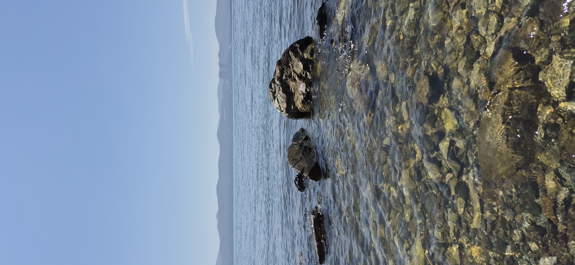 The image depicts a body of water with clear, shallow water revealing a rocky bottom. Several rocks are visible, some partially submerged. In the background, a mountain range stretches across the horizon under a clear blue sky. In the distance, on the far right, a cluster of buildings, possibly a city, can be seen on the coastline. The scene appears to be a natural landscape with some human infrastructure in the distance. This view is consistent with coastal areas of Vladivostok, Russia, particularly those with clear water and a visible mountainous backdrop, such as areas overlooking the Sea of Japan. No people or animals are present in the frame. The lighting suggests it is daytime.