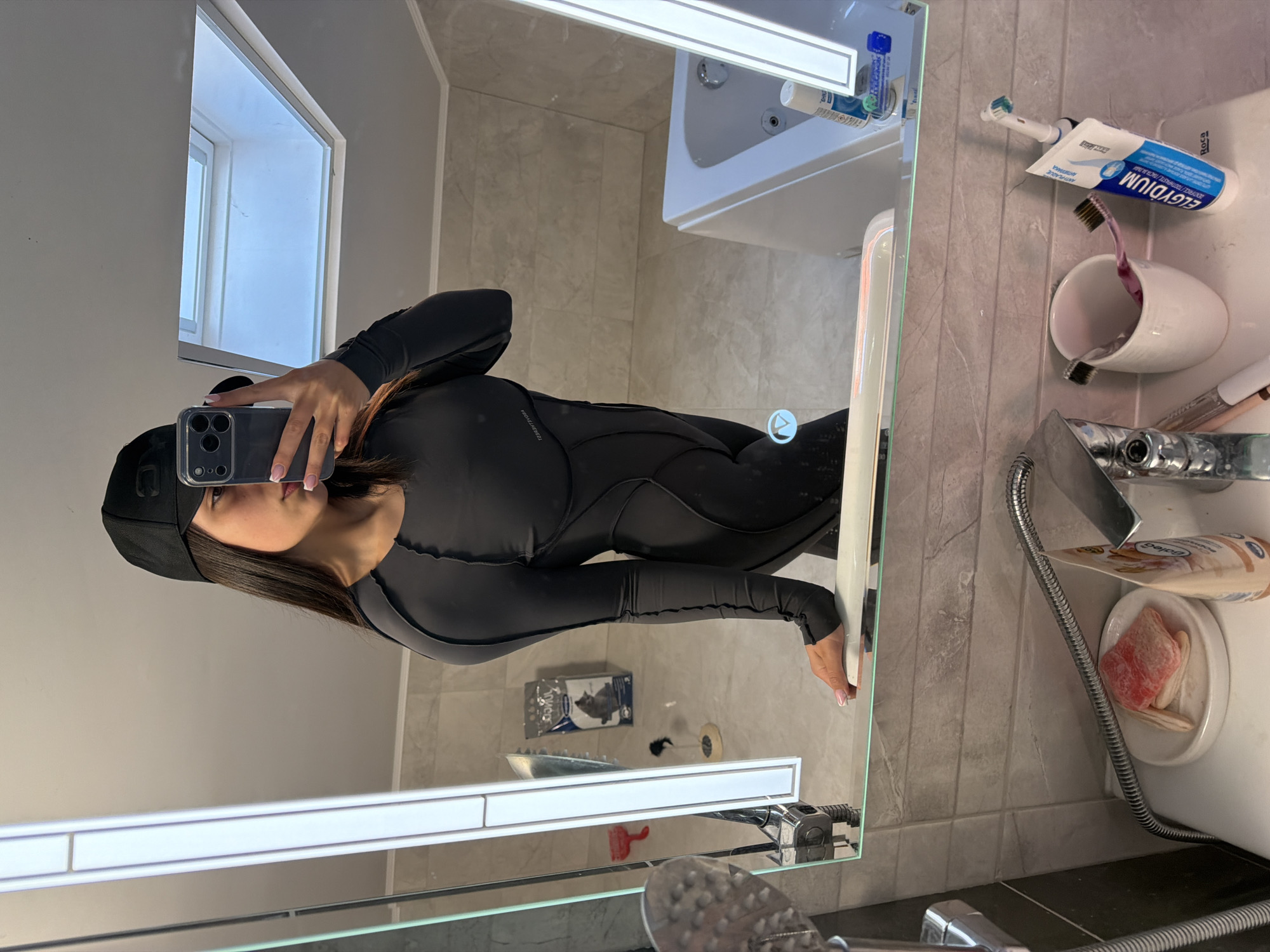 A person wearing a black hat and a black wetsuit is taking a picture of themself in a bathroom mirror. The bathroom has gray tile walls and features a sink, toilet, and bathtub visible in the reflection. Toothbrushes, toothpaste, and other toiletries are placed near the sink. There's a window at the top-left of the reflection. The image lacks specific identifying information to ascertain whether the location is Gabrovo, Bulgaria.
