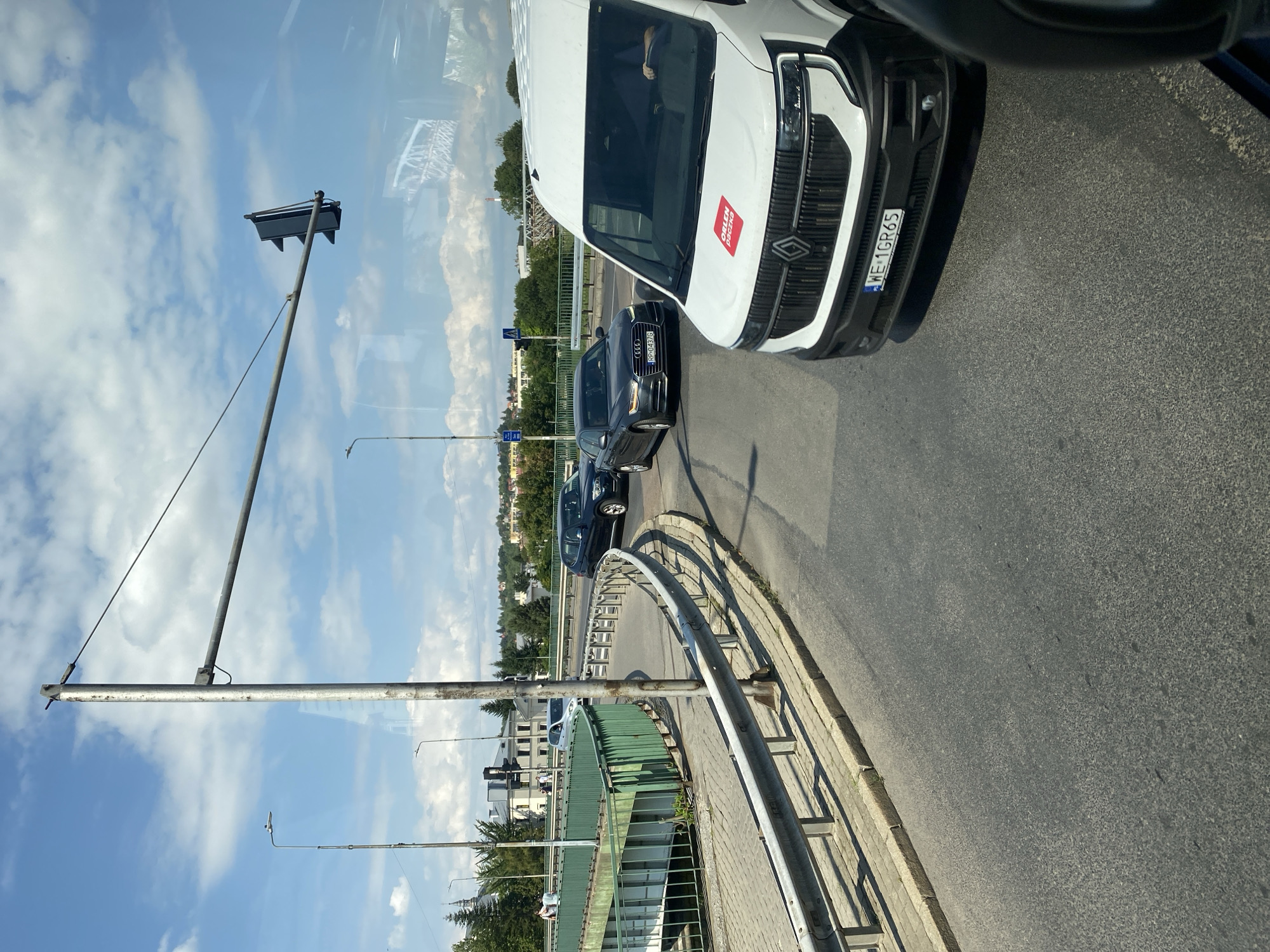 The image captures a view from a vehicle on a road in Przemyśl, Poland, likely on a bridge or overpass, given the elevated perspective and presence of traffic lights and guardrails. In the foreground, a white van with the logo "ORLEN PACZKA" on its hood is visible, and its license plate reads "WE 1GR65". The van is in motion or stopped in traffic. Behind the van, an Audi sedan is seen, its license plate partially obscured but showing "RP-0437G". Further down the road, a dark-colored sedan is also present. The road curves to the left, bordered by a green metal railing on the right side, which is part of an elevated structure. Beyond the railing, more of the elevated structure is visible, with people walking on a paved walkway. In the background, there are buildings, trees, and a blue sky with scattered clouds. A traffic light is suspended over the road. The scene appears to be a typical urban traffic setting.