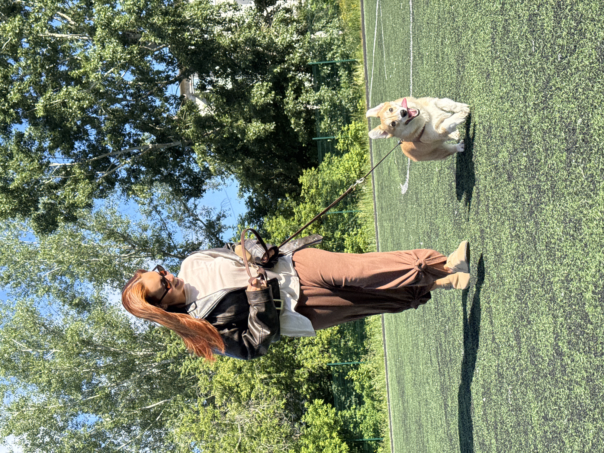 A woman with long, reddish-brown hair and sunglasses is walking a corgi on a leash on a bright, sunny day. She is wearing a black leather jacket over a cream-colored t-shirt and loose, brown pants. The dog is a light brown corgi with its tongue out, appearing to be running or trotting. They are on a green artificial turf field, likely a sports field, with white markings visible. In the background, there are lush green trees and a clear blue sky. The scene suggests a park or recreational area in Chaikovsky, Russia.