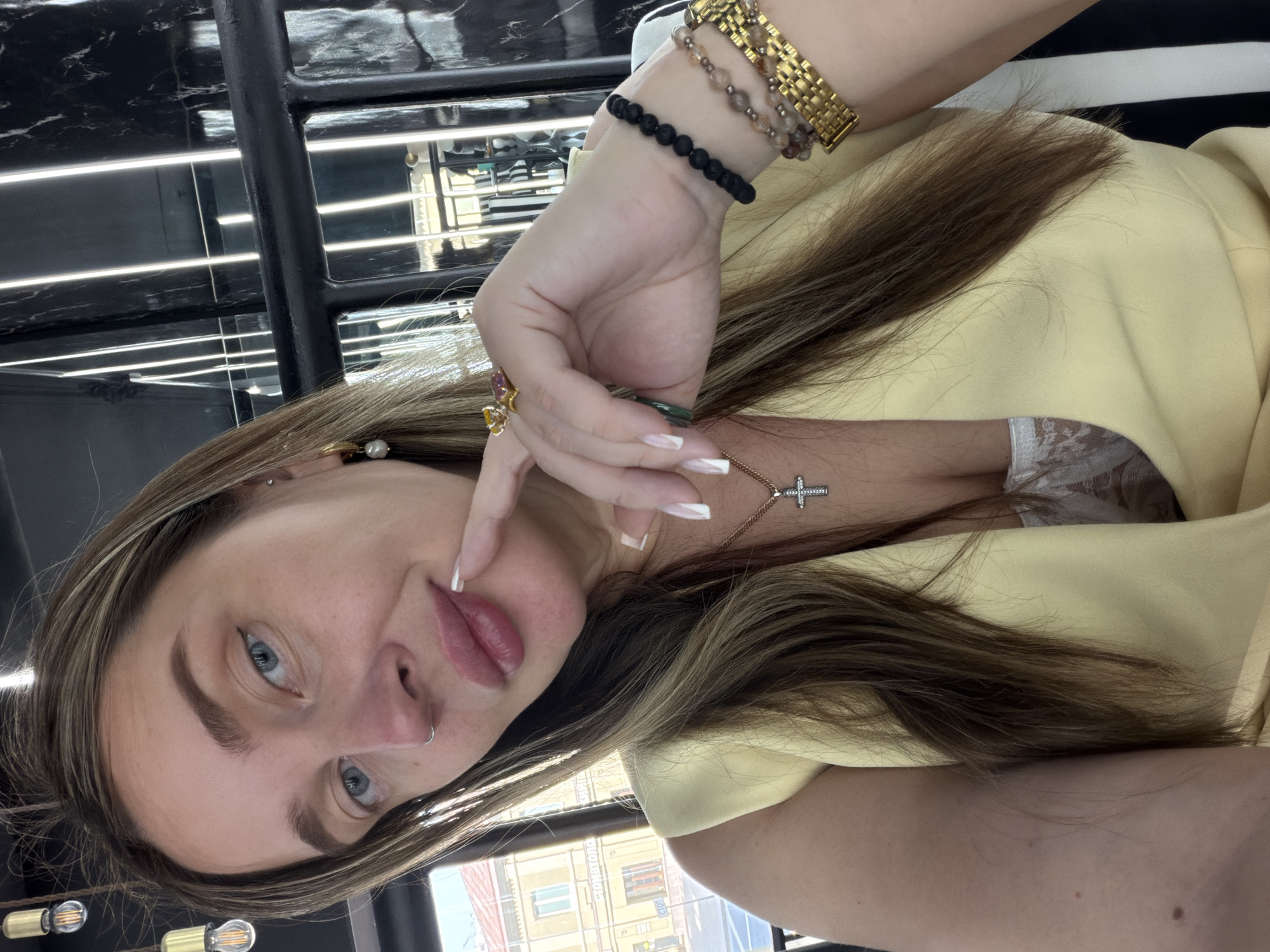 A woman with light brown hair, blue eyes, and a nose ring is posing for a selfie. She has jewelry on her hands and neck, including bracelets and rings. She is wearing a light yellow top. The background suggests a city scene, and based on the architectural style visible through the window, it is Moscow, Russia.