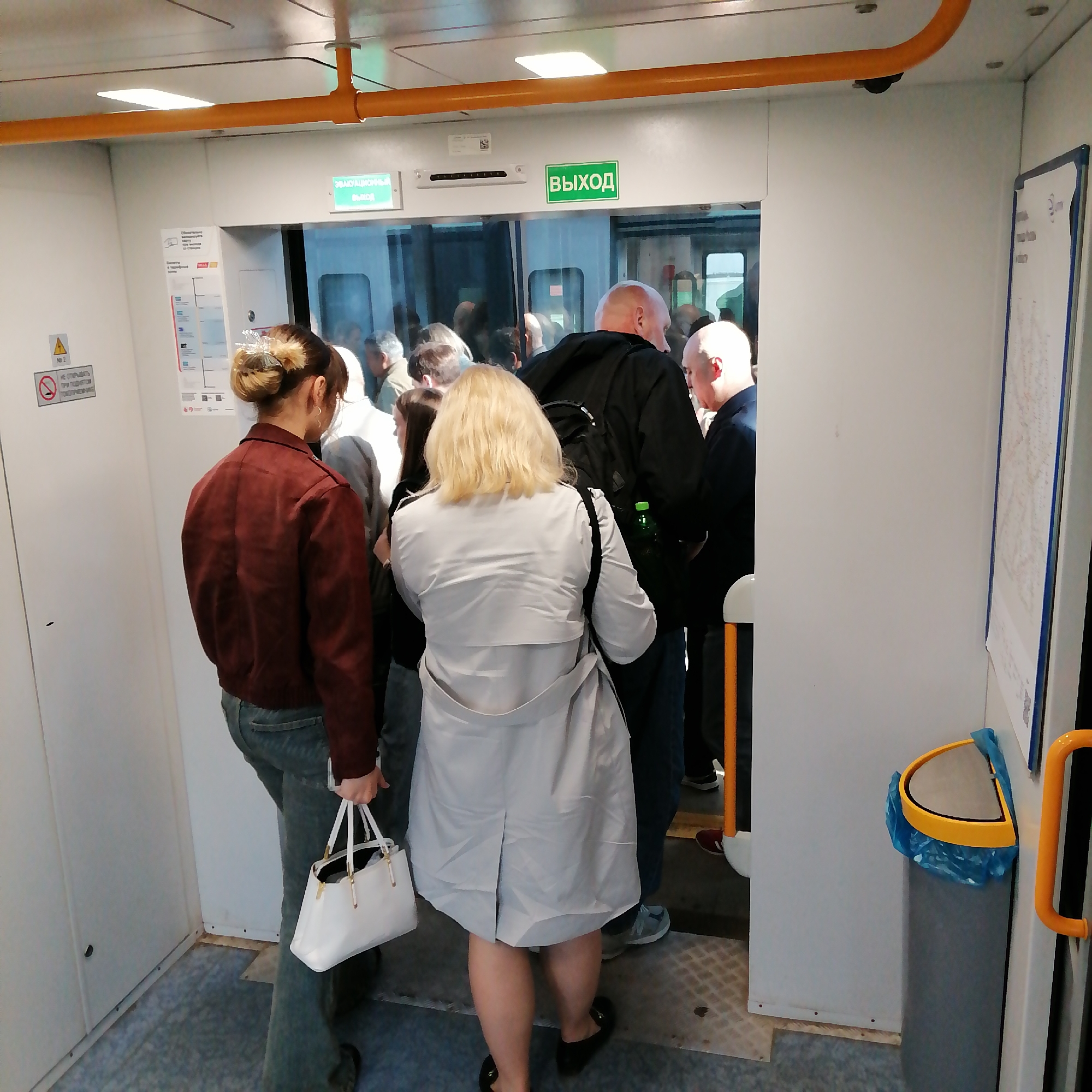 The image shows a group of people on what appears to be a train car. The interior is mostly white with orange handrails. The people are standing near an open doorway, possibly preparing to exit. A woman with blonde hair, wearing a light grey coat, is facing away from the camera in the center of the frame. To her left, a woman with her hair up, wearing a maroon jacket and jeans, holds a white purse. Several other people are visible in the doorway, including a bald man in a black jacket. There is a sign with "Выход" (Exit) in green letters above the doorway. A garbage can with a blue liner is visible on the right side of the frame. The image's location, city, and country are unknown.