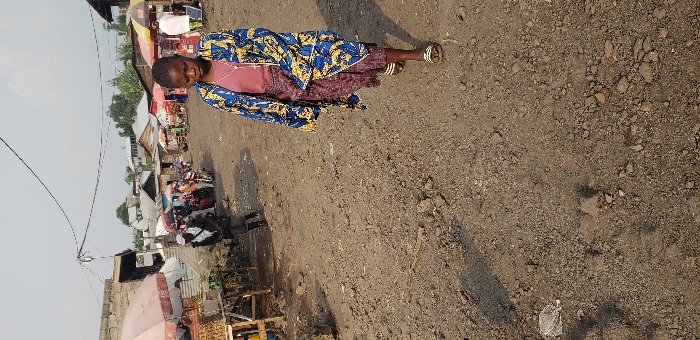 The image depicts a daytime outdoor scene in what appears to be a market or commercial area within Buzove, Democratic Republic of the Congo. The foreground is dominated by a young female, approximately early to mid-teens, walking towards the viewer on an unpaved, uneven dirt and gravel ground surface. She is wearing a long-sleeved, knee-length outer garment with a blue, yellow, and white patterned design over a maroon or reddish-brown top and a patterned maroon or purple skirt. Her feet are shod in white and black striped slide sandals. Her head is slightly bowed, and her gaze is directed downwards.
In the mid-ground and background, an active street or market environment is visible. To the left, a vendor stall features an open pink and white umbrella, with goods displayed on a wooden table. Another individual in dark clothing and a white scarf or wrap is partially visible behind this stall. Further back, numerous people are gathered, and additional market stalls with various colored umbrellas (red, yellow) and structures are present. Several motorcycles or mopeds are parked among the people. Buildings constructed from cinder blocks or brick with corrugated metal roofs line the area. Overhead, multiple electrical wires traverse the scene against a light, overcast sky. The overall lighting suggests natural daylight, with shadows cast on the ground indicating a light source from the right side.