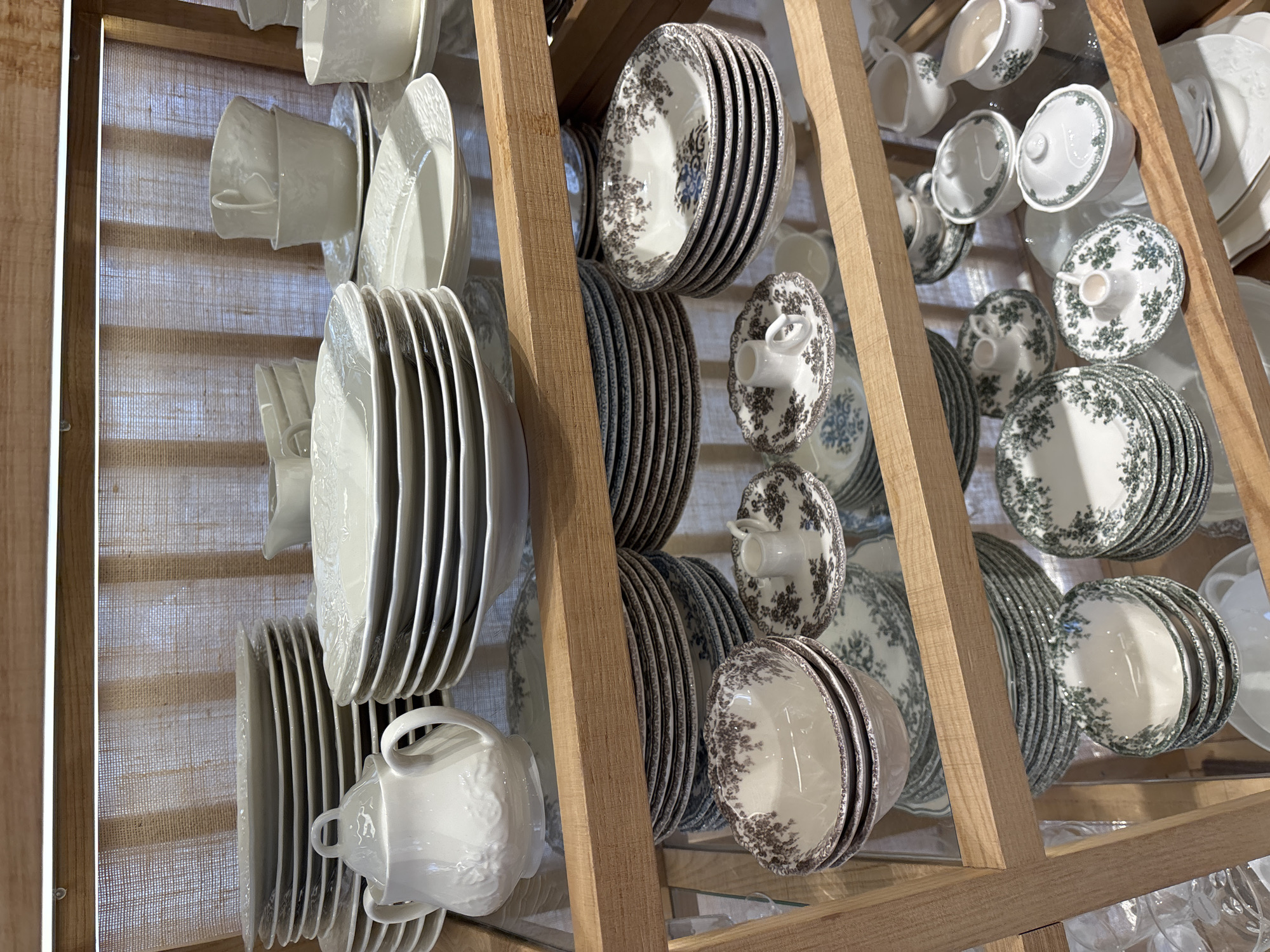 The image shows shelves filled with white and patterned ceramic dinnerware, including plates, bowls, cups, and pitchers. The setting appears to be a retail display, possibly in a kitchenware store or a home goods store. The shelves are made of wood, and the background is a light-colored woven fabric. There are no people visible in the image. The lighting is bright and even, illuminating the details of the dinnerware. The image does not contain any specific geographical markers to indicate its location in Orihuela, Spain.