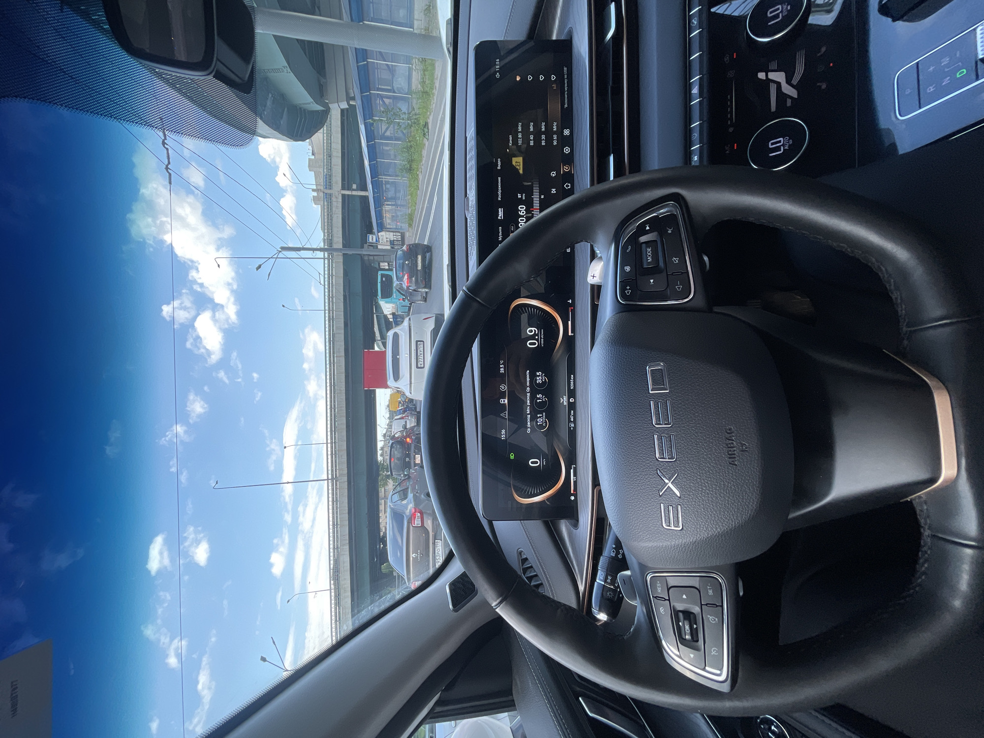 The image shows the interior of a car, likely in traffic. The view is from the driver's perspective, looking through the windshield and at the dashboard. The dashboard features a digital display showing various information, including speed, fuel consumption, and radio stations. The steering wheel is visible, with controls for media and cruise control. The exterior scene visible through the windshield suggests a city or urban environment with elevated structures and other vehicles on the road. The sky is blue with some clouds, indicating daytime. The time displayed on the dashboard is 15:56. Some visible text includes "EXCEED" on the steering wheel, license plate numbers like "H499BT877" and "953PO 198", and various labels on the digital display related to car functions and media. There are no people clearly visible in the image. The overall impression is one of a modern car interior in a typical urban traffic situation during the afternoon.