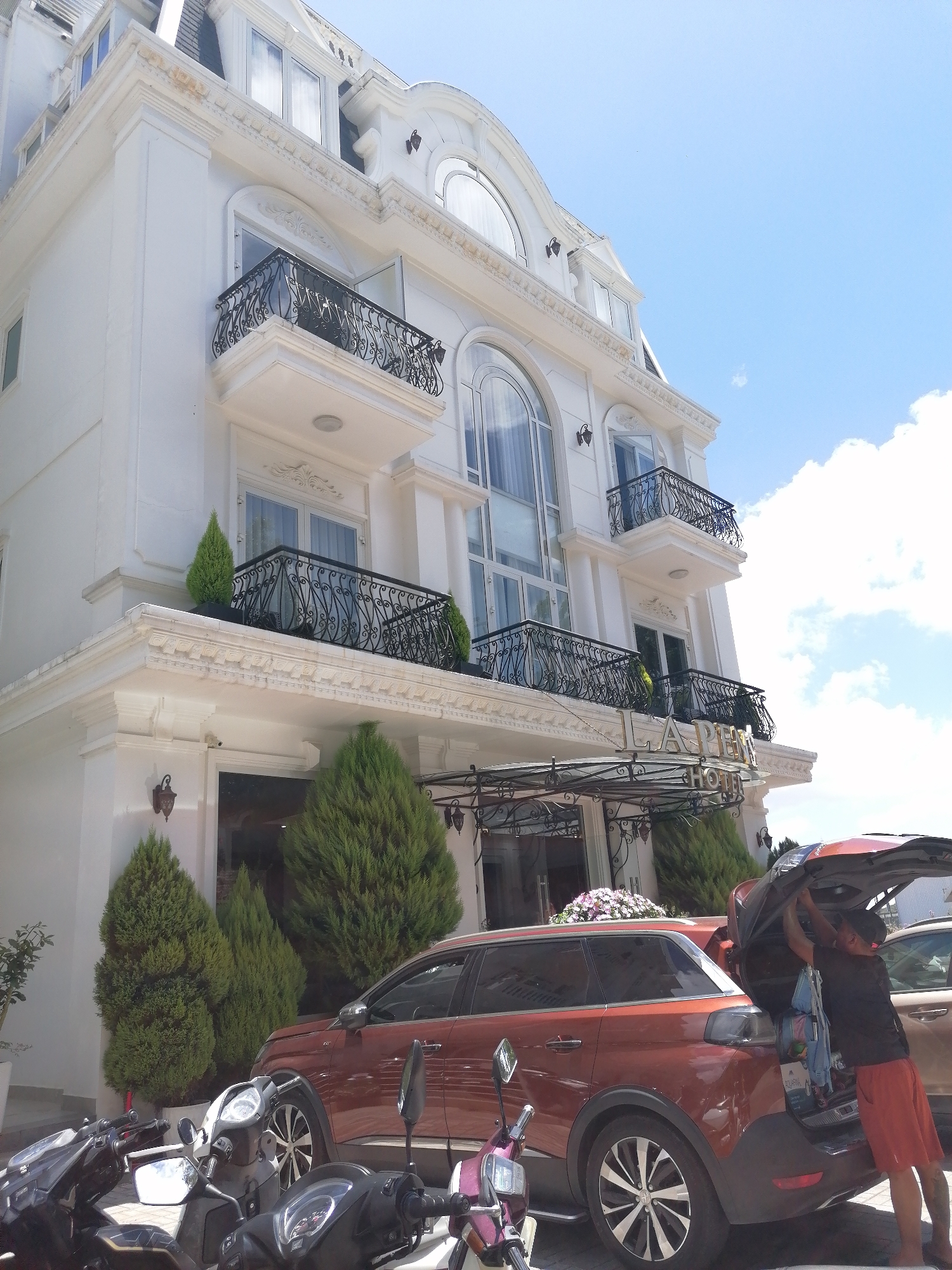 A man is loading items into the open trunk of a red SUV parked in front of a white, multi-story hotel with ornate balconies. The hotel is identified by a sign that reads "LAPEN HOTEL". Several motorcycles are parked to the left of the SUV, and decorative cypress trees are planted in front of the hotel entrance. The scene appears to be set in Da Lat, Vietnam, as indicated by the architecture and vegetation typical of the city. The sky is clear with scattered clouds.