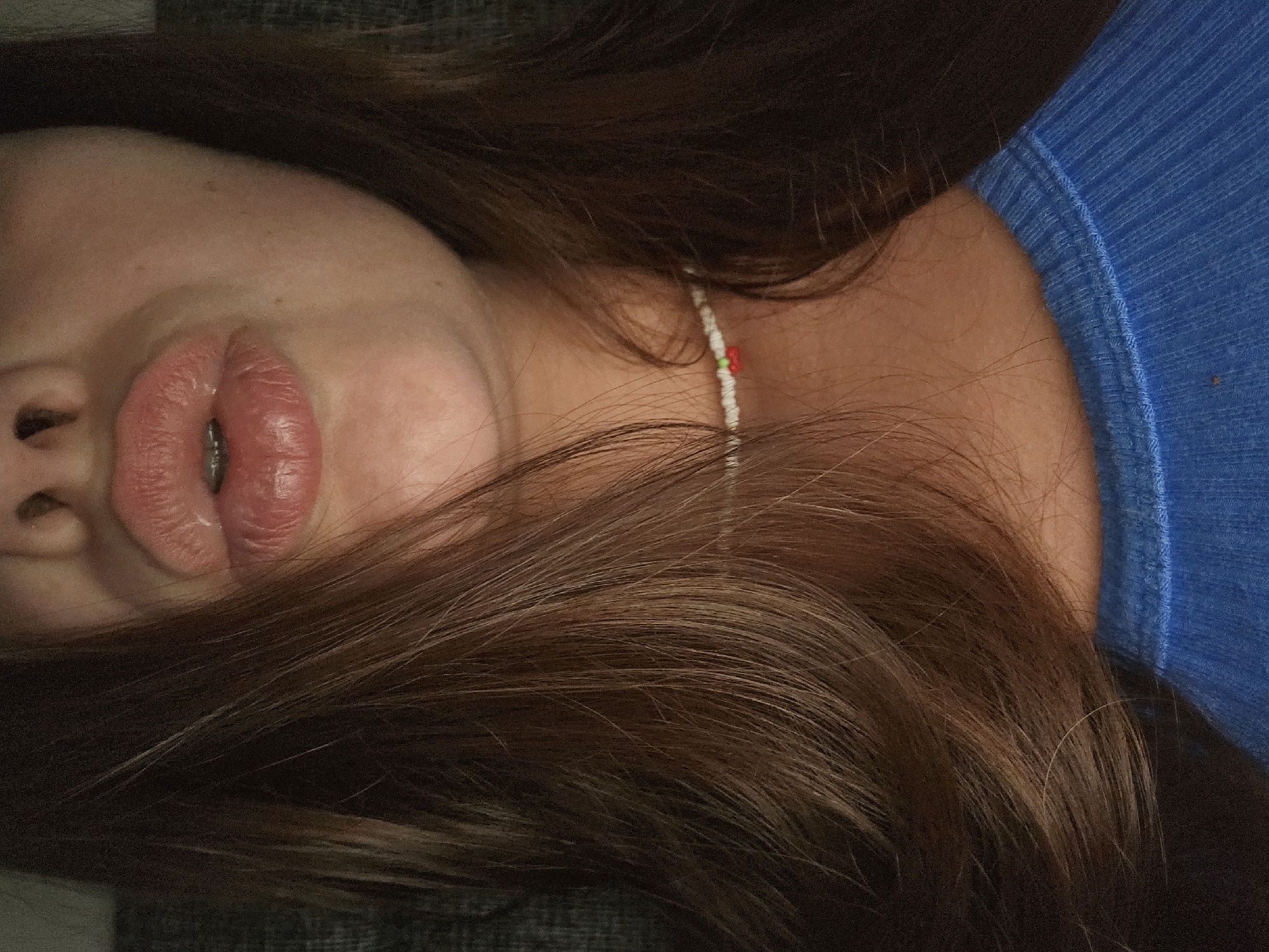 A close-up, chest-up shot focuses on a person's face, with their mouth pursed into a kiss. Their lips are full and have a glossy sheen, and braces are visible on their teeth. Long, dark brown hair frames the face, falling across the left side of the image. The person is wearing a blue, ribbed t-shirt, and a delicate beaded necklace with small red and green beads is visible on their neck. The background is dark and out of focus, providing no discernible environmental details. The image was taken in Petropavlivska Borshchahivka, Ukraine.