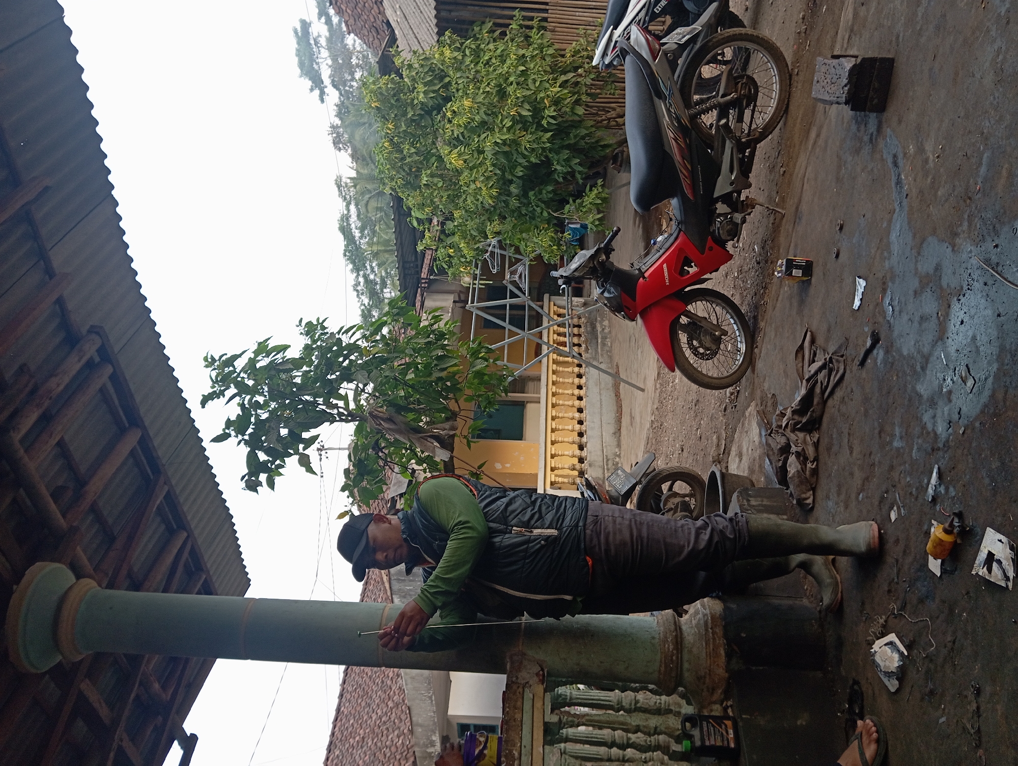 In Kudus, Indonesia, a man wearing a black cap, a green long-sleeved shirt, and a dark vest stands under a covered structure, holding a metal rod and seemingly preparing to insert it into a vertical post. He wears dark pants and tall green boots. The ground is concrete and appears to be a workshop or outdoor area, with scattered debris, including fabric scraps, plastic bottles, and metal pieces. A red motorcycle is parked to the right, and another is partially visible behind it. In the background, houses with red tile roofs and lush green trees are visible, suggesting a residential or semi-rural setting. The sky is overcast.