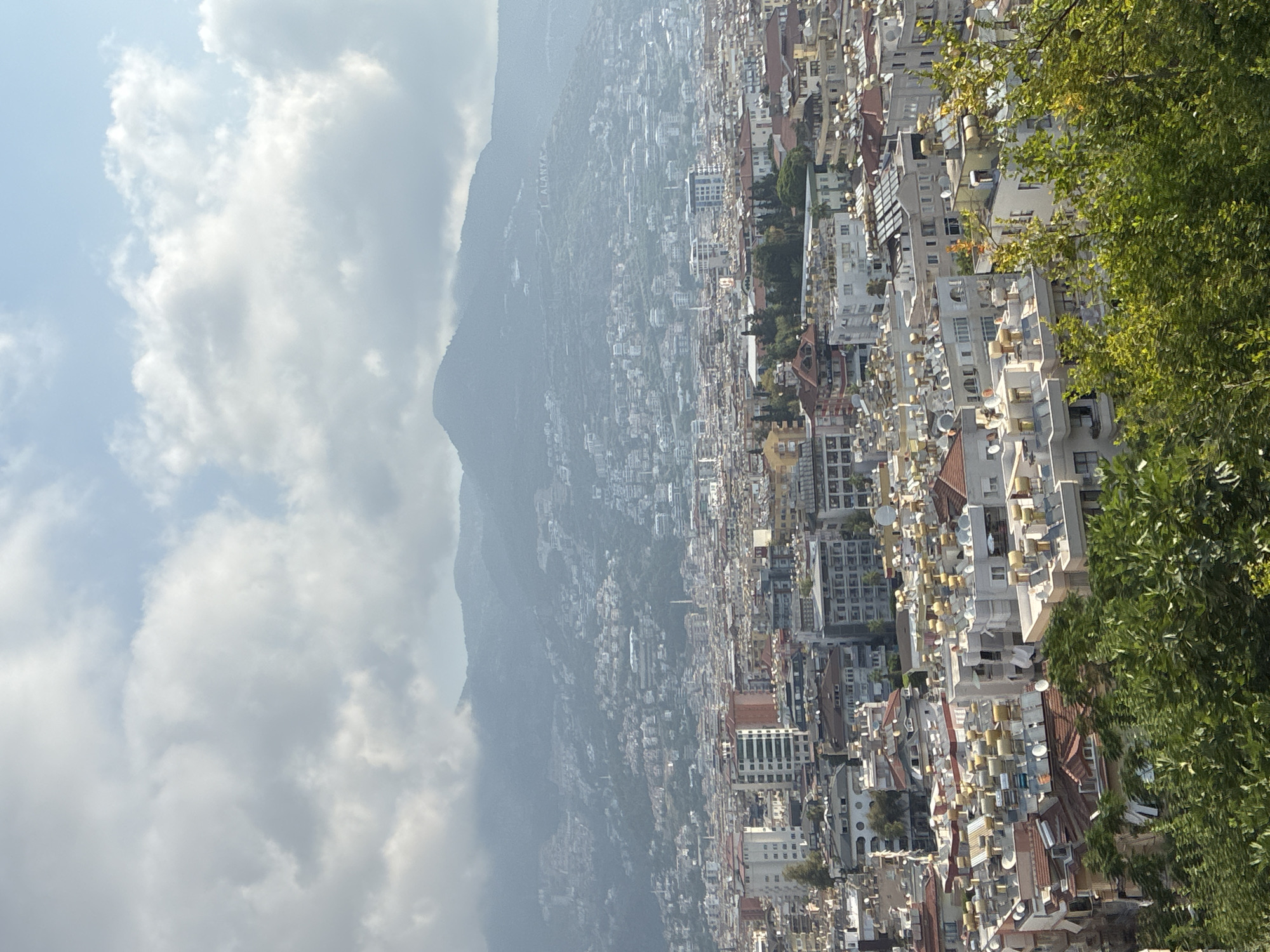 This is a high-angle, vertical, wide shot of the city of Alanya, Turkey, taken from a viewpoint above the city looking towards a large mountain range in the background. The foreground is filled with the dense green foliage of trees and bushes. Below the foliage, the midground and background are dominated by a sprawling urban landscape of buildings. Numerous buildings, varying in height and architectural style, are packed closely together. Many of the rooftops are covered with arrays of solar water heaters, giving them a distinctive appearance. A prominent feature in the far background, on the mountain slope, is a large sign that reads "I ALANYA" in white letters. The sky above is partly cloudy with scattered white and gray clouds against a light blue sky. The overall scene depicts a densely populated coastal city nestled against a mountainous backdrop.