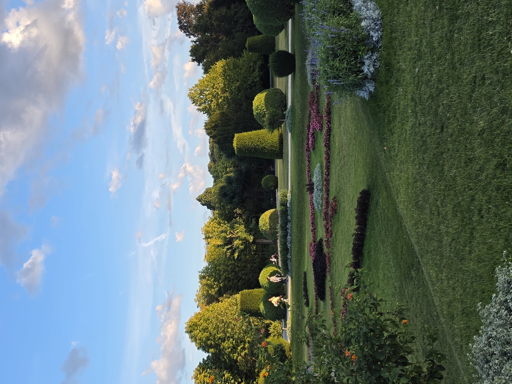 The image depicts a scenic view of the Schönbrunn Palace gardens in Vienna, Austria, under a bright blue sky with scattered white clouds. The foreground features a meticulously manicured lawn with flowerbeds containing a variety of colorful flowers in shades of pink, red, and purple, as well as a large shrub with purple flowers on the right. In the middle ground, several individuals are walking along a paved pathway that winds through the park. These individuals appear to be casually strolling and enjoying the surroundings.
The background is dominated by a dense line of mature trees, their lush green foliage creating a natural backdrop. Interspersed among the trees and on the lawn are various topiaries, sculpted into elegant geometric shapes such as cones and spheres, showcasing the park's formal garden design. Sunlight dapples the scene, highlighting the vibrant greens of the grass and trees and casting soft shadows. The overall impression is one of a serene and well-maintained public garden, inviting visitors to relax and appreciate the natural beauty and horticultural artistry.