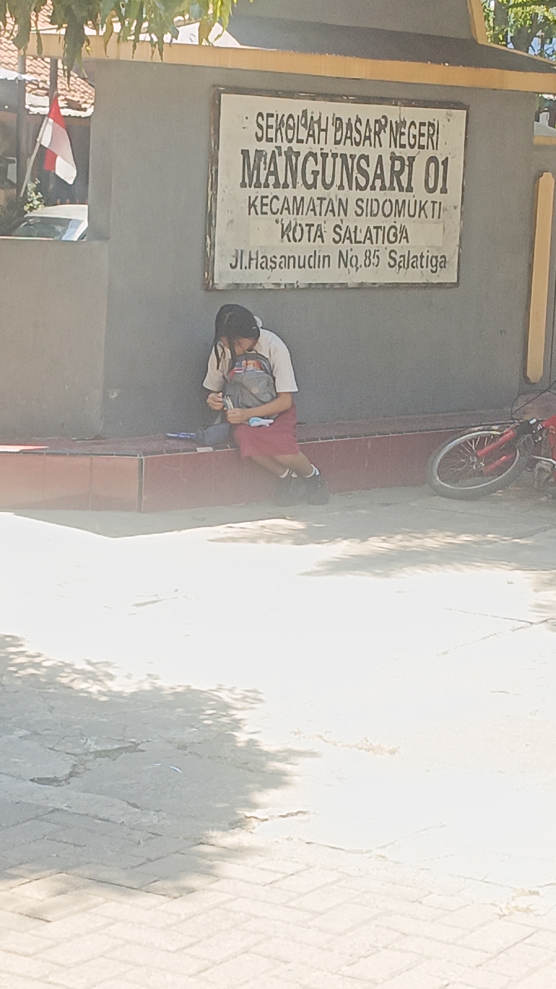 The image shows a sign in front of a school in Salatiga, Indonesia. The sign reads "SEKOLAH DASAR NEGERI MANGUNSARI 01 KECAMATAN SIDOMUKTI KOTA SALATIGA Jl. Hasanudin No. 85 Salatiga". A young student wearing a school uniform sits on a low wall in front of the sign, appearing to be looking at something in their hands. A backpack is next to them. A red bicycle is parked to the right of the student. The scene is outdoors with sunlight casting shadows on the ground.