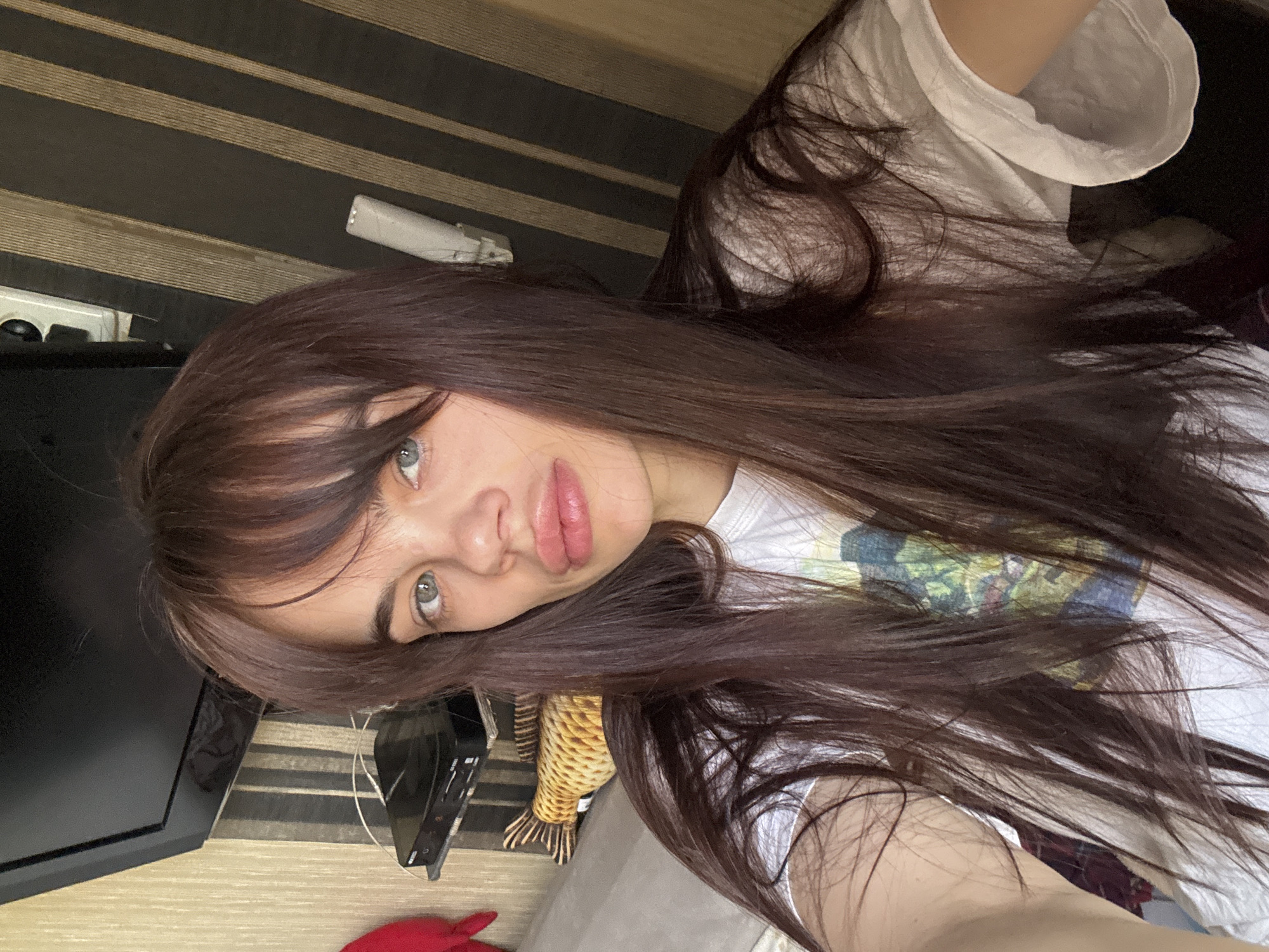 A young woman with long brown hair and bangs is taking a selfie. She is wearing a white t-shirt. Her eyes are light-colored, and she has full lips. In the background, there is a striped wall and a television. There is also what appears to be a plush fish on a grey couch. The location is likely in an indoor setting. There is no evidence to support the photo was taken in the city of Rostov-on-Don, Russia.