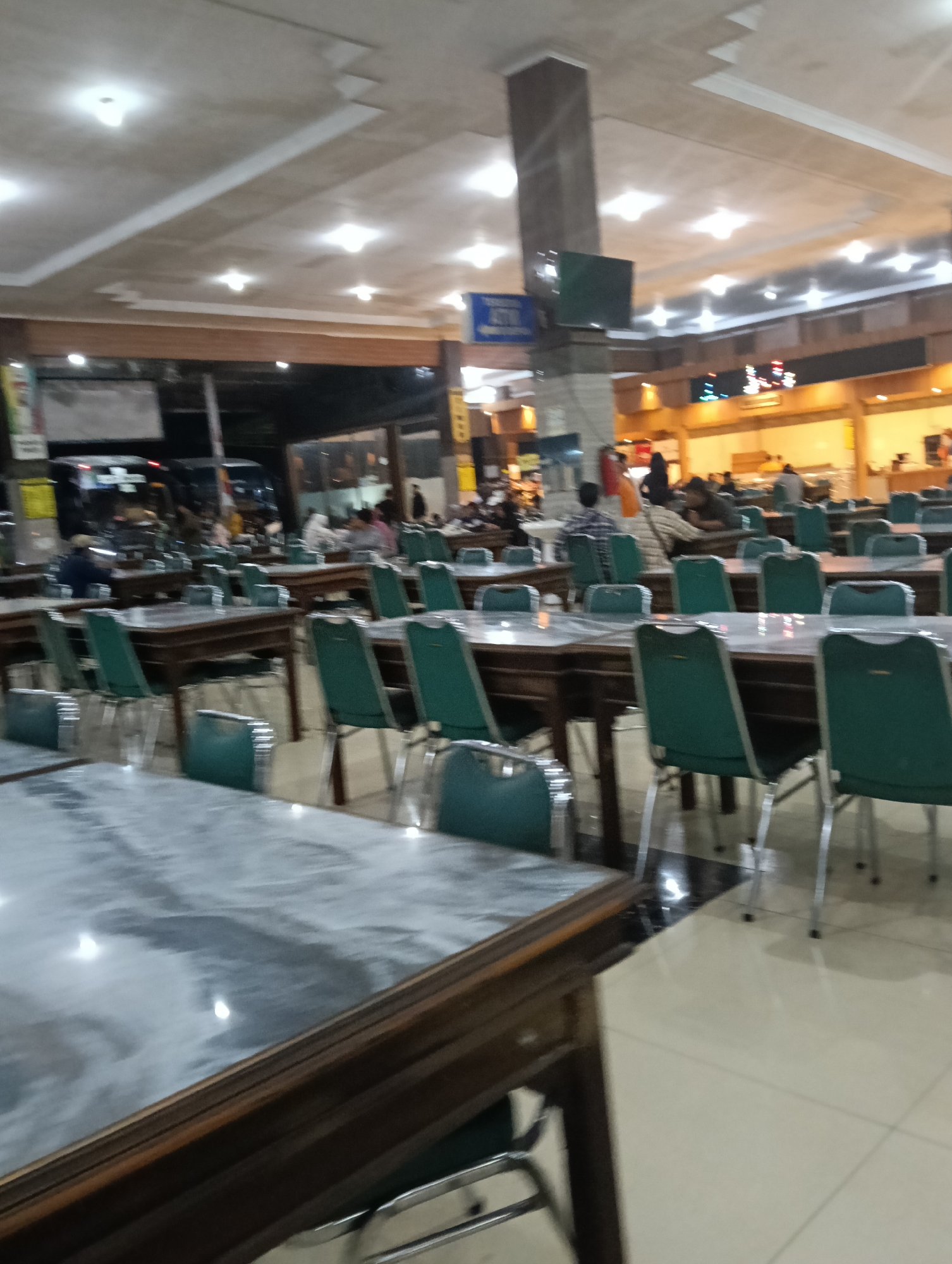 The image depicts a spacious indoor dining area with numerous tables and chairs. The tables have dark wood frames and light-colored, possibly marble or laminate, tops. The chairs are predominantly green with silver metal legs. The dining area is illuminated by ceiling lights, some of which are recessed. In the background, there are more tables and chairs, as well as a glimpse of people dining or conversing. The overall impression is that of a busy restaurant or canteen. The environment appears clean and well-maintained. The perspective suggests the photograph was taken from a seated position at one of the tables. There is no indication of the specific time of day, though the artificial lighting implies it could be evening or indoors during daylight. There is no visible signage or architectural feature that specifically identifies the location as Cikawung, Indonesia.