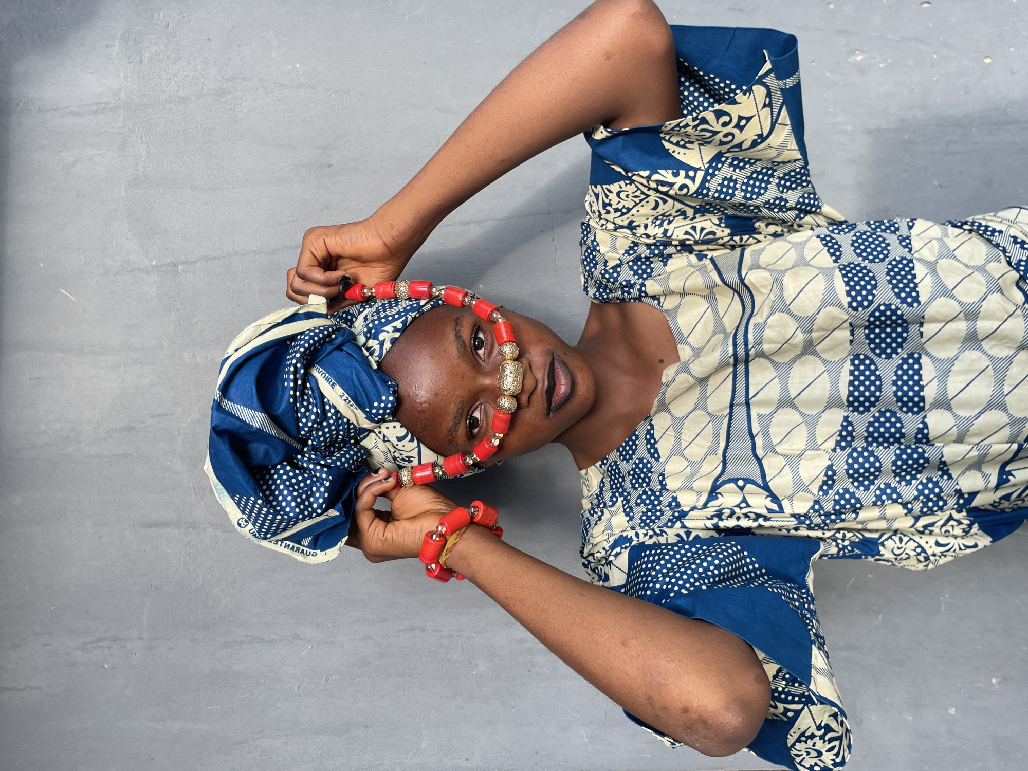 A woman is lying on a gray surface. She wears a blue and white patterned dress and a matching headwrap. She has a red beaded necklace and a matching bracelet. A decorative gold nose ring is visible. The location is not identifiable as Badagry, Nigeria based solely on the visual information.