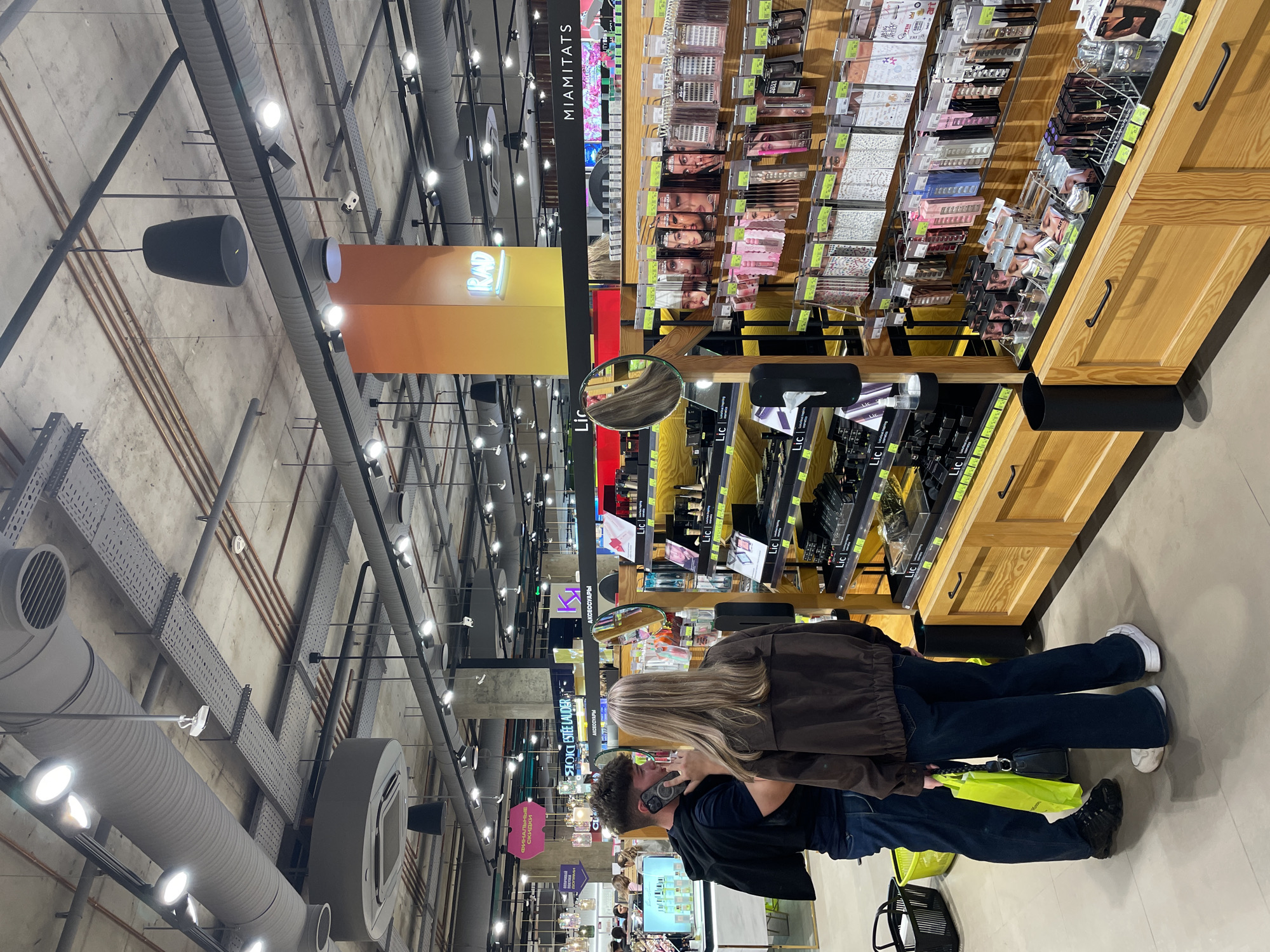 Two individuals, a man and a woman, are standing in a store aisle, potentially at a cosmetics or beauty supply section. The woman has long blonde hair and is wearing a brown jacket and blue jeans. The man has dark curly hair and is wearing a black shirt and blue jeans. He appears to be holding a phone to his ear. They are near a display with various makeup and cosmetic products, which are neatly arranged on shelves. A yellow bag is visible at their feet, and a black shopping basket is nearby. The store has high ceilings with visible industrial ductwork, pipes, and light fixtures. Signage for various brands and promotions is visible. Based on the signs "MIAMITATS" and "RAD" and other store signs, this image was captured in the Golden Apple (Zolotoe Yabloko) store at Aviapark, Moscow, Russia.