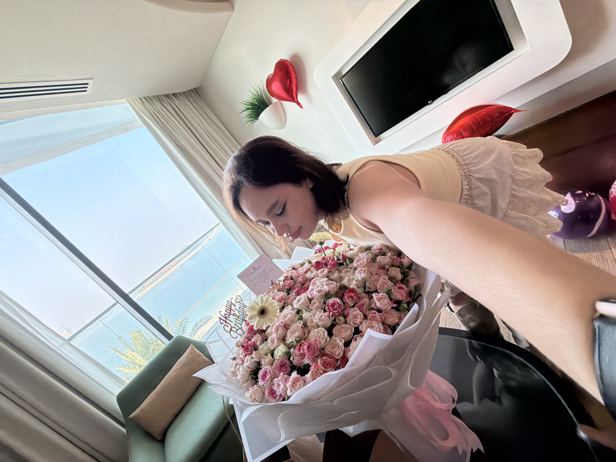A woman is shown in an interior room, possibly a hotel room, with a large bouquet of pink and white roses. She is leaning over the bouquet, appearing to smell or examine it. The room has a large window overlooking a body of water, likely the Arabian Gulf, with palm trees and buildings visible in the distance, suggesting a coastal location within Dubai, United Arab Emirates. Red heart-shaped balloons are visible on the wall and shelf. A flat-screen television is mounted on the wall above a shelf. The woman is wearing a light-colored sleeveless top and a white ruffled skirt. A green armchair with a tan pillow is visible near the window. The scene suggests a celebratory occasion, possibly a birthday.