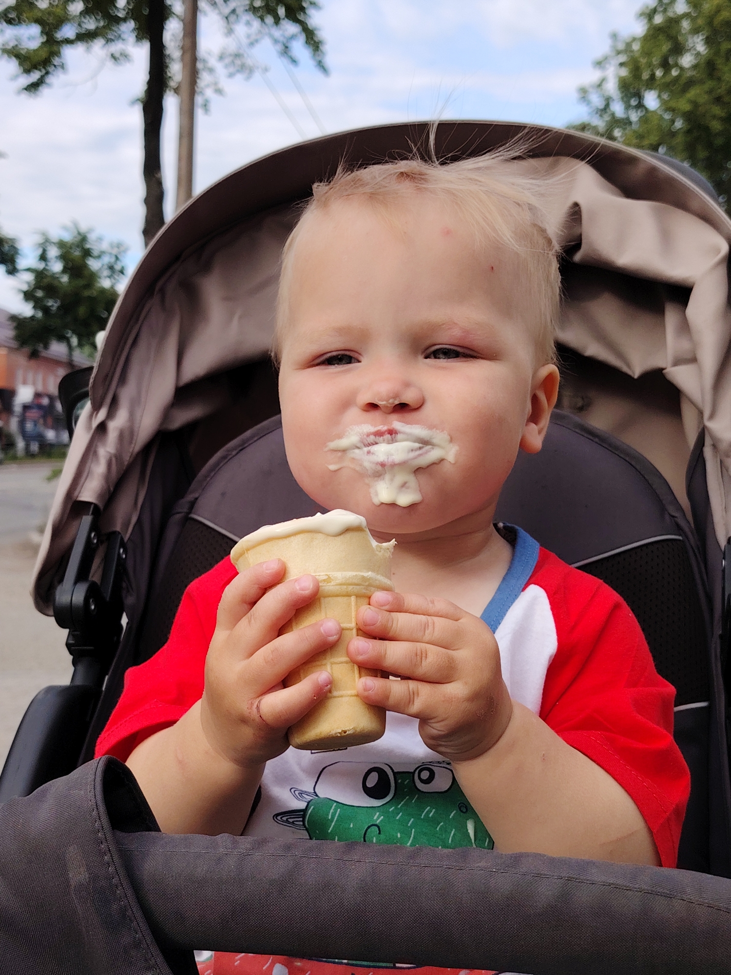 A blonde-haired toddler, approximately 1-2 years old, is depicted in a medium close-up, seated in a dark grey and beige baby stroller in an outdoor urban environment within Chaikovsky, Russia. The child, wearing a red and white short-sleeved t-shirt with a partially visible green graphic on the chest, holds a waffle ice cream cone containing white ice cream with both hands. White, melted ice cream covers the child's mouth and nose. A small red abrasion is visible on the child's forehead above the left eyebrow, and a reddish swelling is present directly below the left eye. A faint, curved scratch mark is observed on the inner surface of the child's right wrist. The background features blurred green foliage, a utility pole, and distant structures under an overcast sky, suggesting a street or park setting.