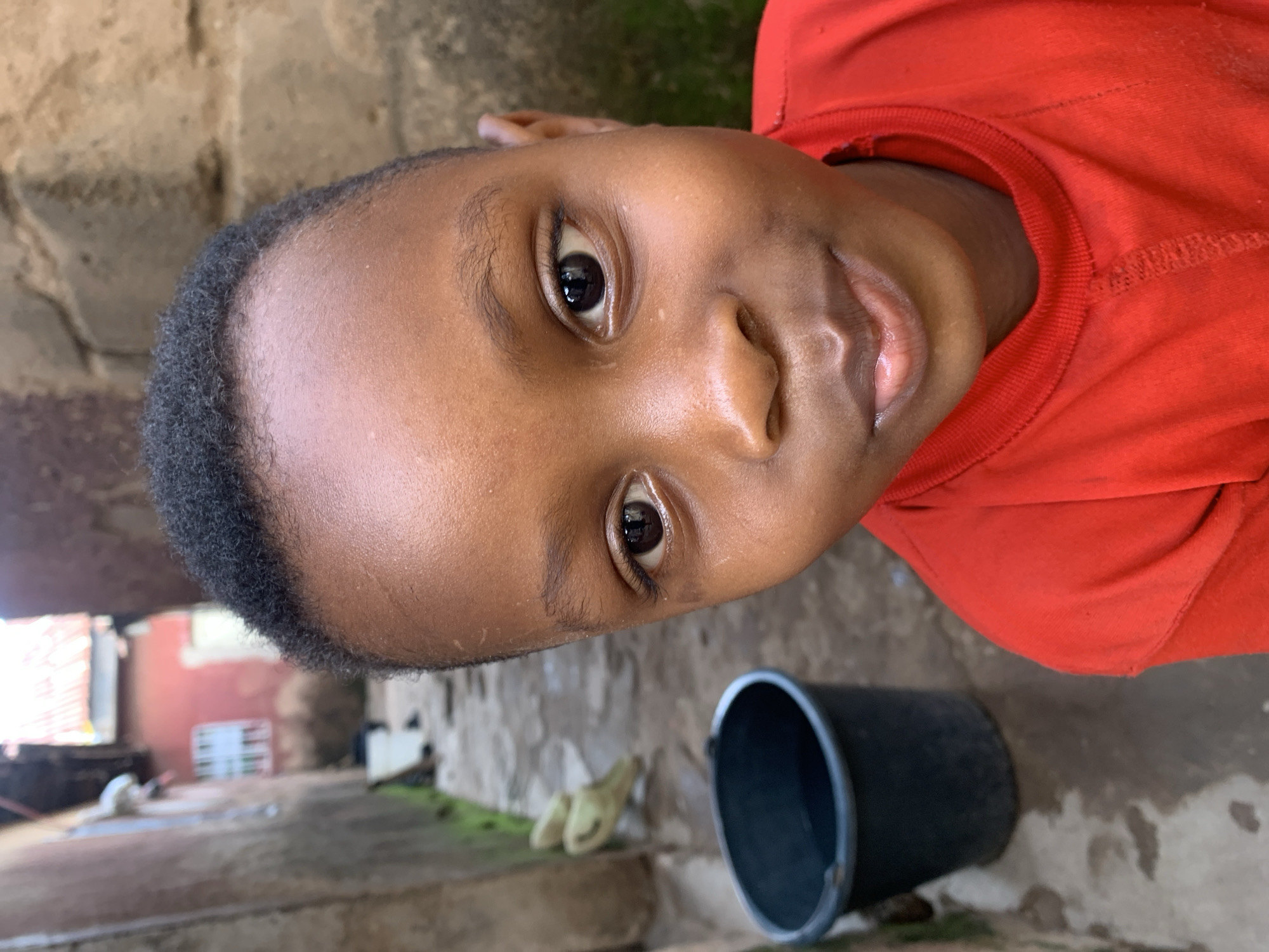 The image features a close-up, rotated shot of a young person wearing a red t-shirt. The subject's face is the primary focus, with dark skin and brown eyes. Their hair is short and dark. In the background, a rough, textured wall made of what appears to be stone or concrete is visible. To the right of the subject, a black bucket is partially visible, with its rim closest to the camera. The ground appears to be dirt or concrete, with some patchy green growth. In the further background, hints of a building and some other objects are discernible, suggesting an outdoor or semi-outdoor setting, possibly a courtyard or alleyway in a residential area. The lighting suggests it could be daytime, but there are no clear indicators of weather or specific time of day. The person's expression is neutral. There is no visible text in the image. The setting context is Jos, Nigeria.