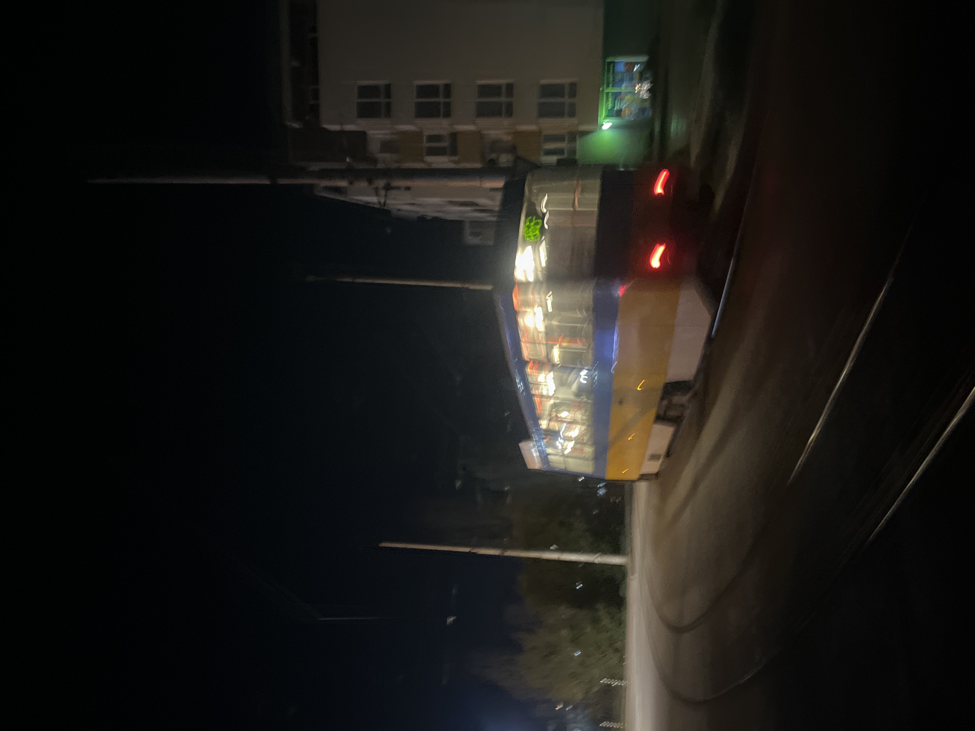 A long, yellow bus is parked on a street at night in Sofia, Bulgaria. The bus is illuminated, showing its interior through its windows. A building with multiple windows is visible behind the bus. The road is dark, and minimal ambient lighting is present beyond the immediate bus and building area.