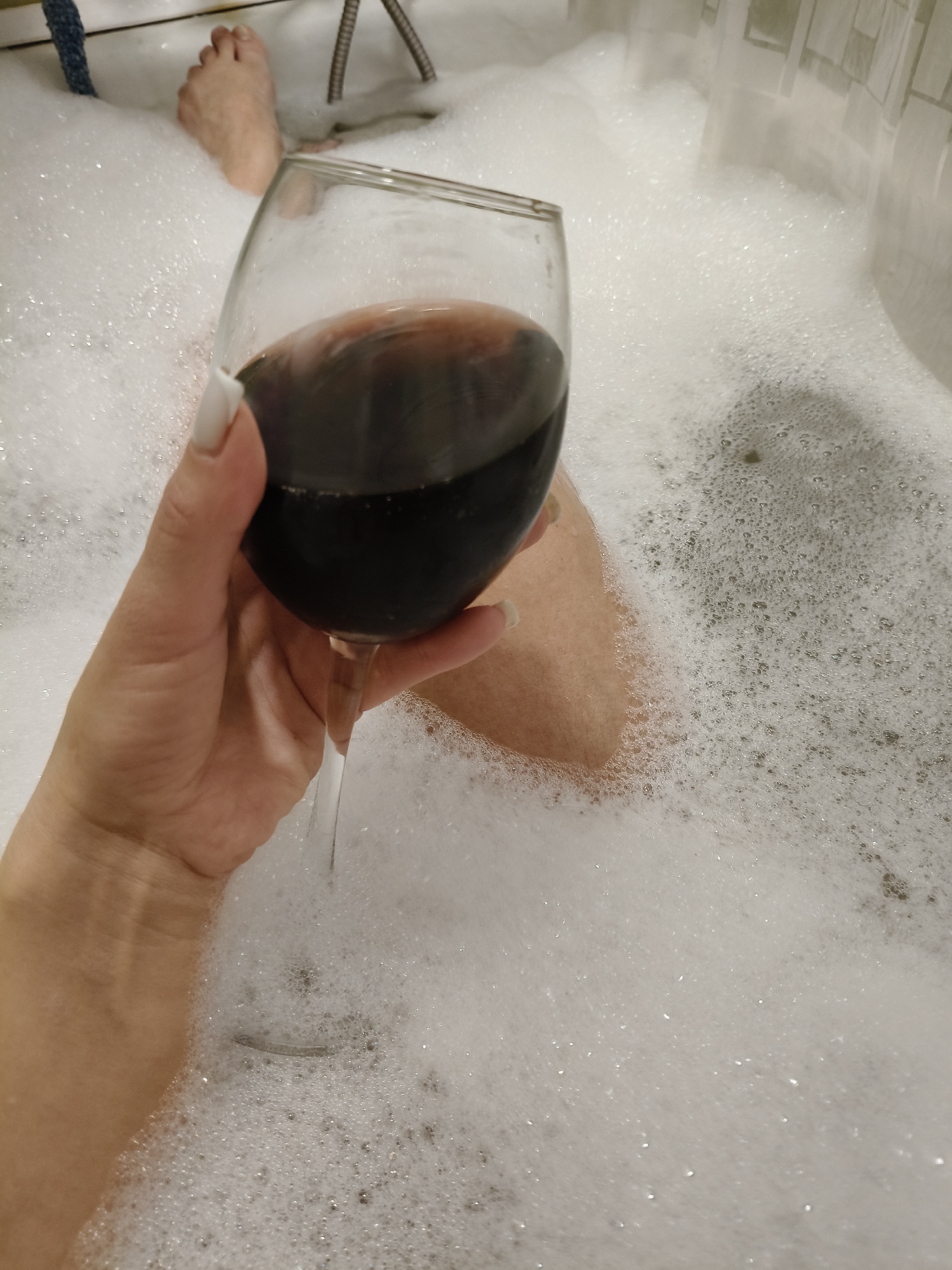 The media file presents a first-person perspective of a person taking a bubble bath in Chaikovsky, Russia. The central focus is a hand, likely female, with neatly manicured white fingernails, holding a stemmed wine glass containing a dark red liquid, presumably red wine.
The bathtub is filled with a thick layer of white, frothy bubbles, almost entirely obscuring the water beneath. Partially visible in the bubbly water are the person's feet in the upper left corner and a portion of their leg or thigh beneath the wine glass. A shower hose or fixture is visible in the upper right background, along with what appears to be a tiled wall or a shower curtain.
The scene conveys an atmosphere of relaxation and personal indulgence. The lighting is artificial, typical of an indoor bathroom setting, suggesting an evening or private unwinding moment. There is no visible text within the image.