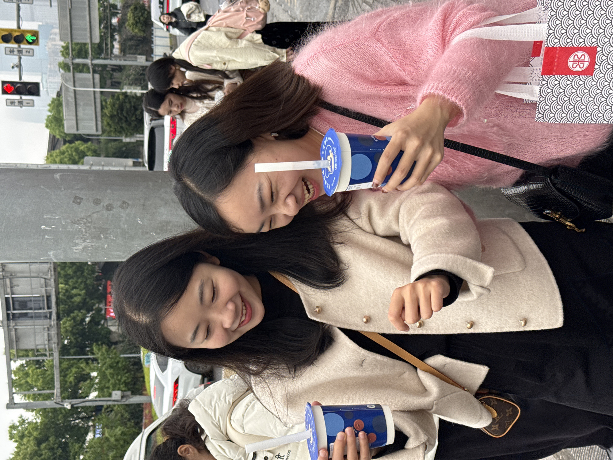 This image captures a street scene in Nan'an District, China, under an overcast sky. In the foreground, two young East Asian females are shown from the chest up, engaged in a positive interaction. The female on the left wears a cream-colored collared jacket with gold buttons over a black top and black pants. A small brown shoulder bag with a visible "LV" monogram hangs from her right shoulder. She holds a blue and white beverage cup with a white straw in her left hand and gestures with her right hand, smiling as she looks towards the right. The female on the right wears a fuzzy pink sweater. A black crocodile-patterned shoulder bag is visible on her left side. She holds an identical blue and white beverage cup with a white straw in her right hand and a white and black patterned shopping bag with a red "SSS" logo in her left hand. She is actively laughing, looking towards the left.
Separating them slightly is a large gray metal pole. In the mid-ground to the left, the back of another East Asian female in a white puffer jacket is partially visible. Further in the mid-ground to the right, two other East Asian females are visible, one in a light-colored top and another in a cream jacket with a pink backpack, appearing to converse. In the far background right, another East Asian female in a dark jacket is seen walking. The background features a city street environment with white vehicles, including a van on the far left and cars on the street. Traffic lights are visible, with a red circular light and a green arrow light on an overhead gantry displaying Chinese characters. Green trees line the street, and tall buildings are visible in the distant background. The ground surface appears to be paved.
