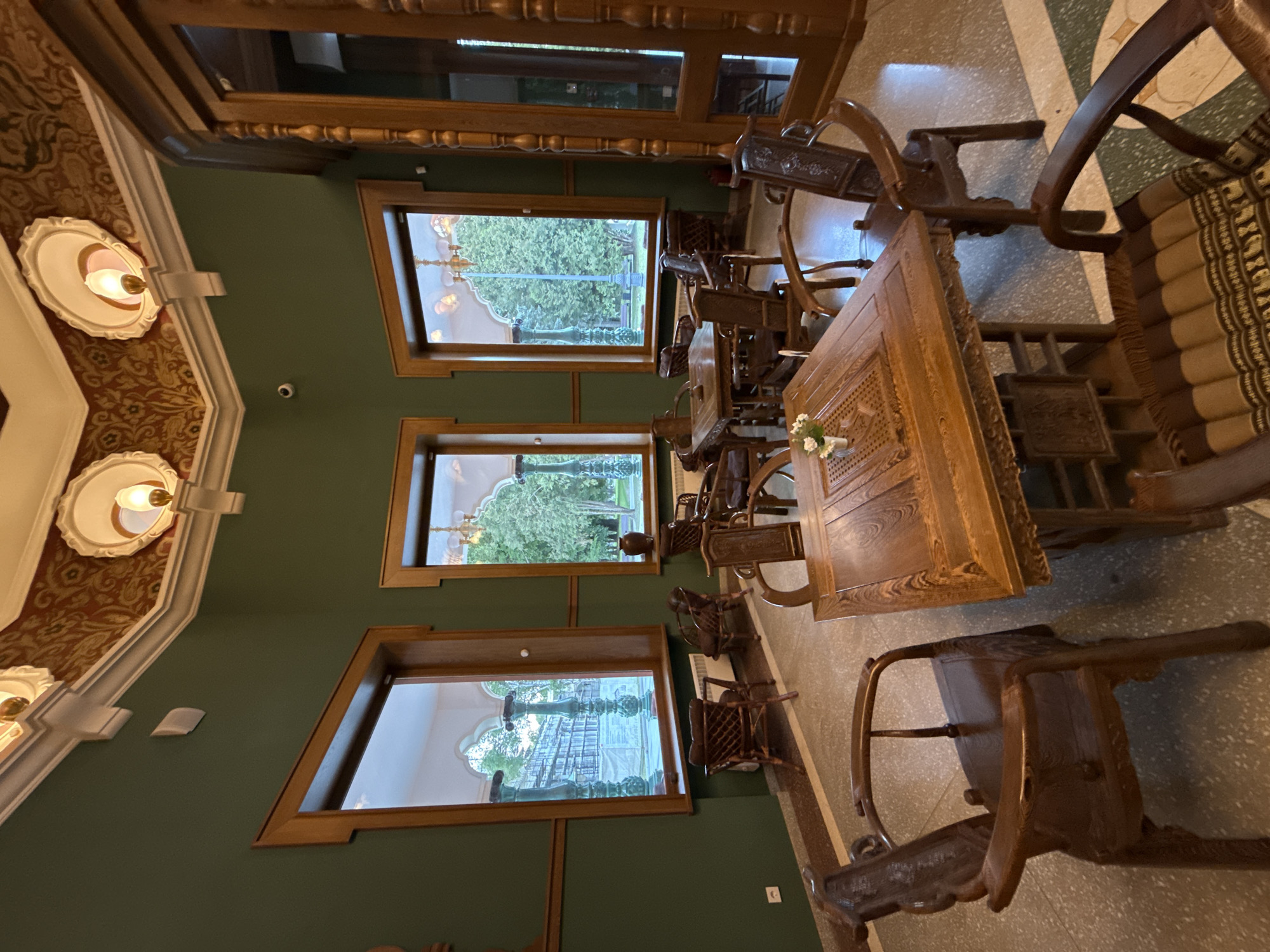 The image displays the interior of an elegantly furnished room, likely a cafe, restaurant, or a common area within a historic building in Moscow, Russia. The room is characterized by a traditional and ornate design.
The walls are painted a deep green, contrasting with decorative white molding and a patterned wallpaper or paneling in shades of brown and orange/red on the upper sections and ceiling. Several dark wooden tables and chairs are arranged throughout the space. The furniture is intricately carved, with some chairs featuring detailed backrests and upholstered seats, one of which has a patterned fabric cushion. A prominent wooden table in the mid-ground holds a small vase containing white flowers.
Multiple large windows with dark wooden frames offer views of lush green trees and outdoor greenery, suggesting a garden or park setting outside. The windowpanes reflect parts of the interior ceiling and possibly architectural details from the exterior. The windows themselves appear to have custom or uniquely shaped frames, hinted at by their reflections.
The floor is covered with patterned tiles featuring geometric designs in light and dark hues. Ornate, classic-style wall-mounted light fixtures with exposed bulbs are visible on the upper walls. A small white security camera is mounted on the green wall, along with another small white wall-mounted device.
The room is devoid of people, appearing tranquil and set up for guests. The natural light filtering through the windows suggests it is daytime. No visible text is present in the image. The overall impression is one of classic elegance and a rich, historical atmosphere.