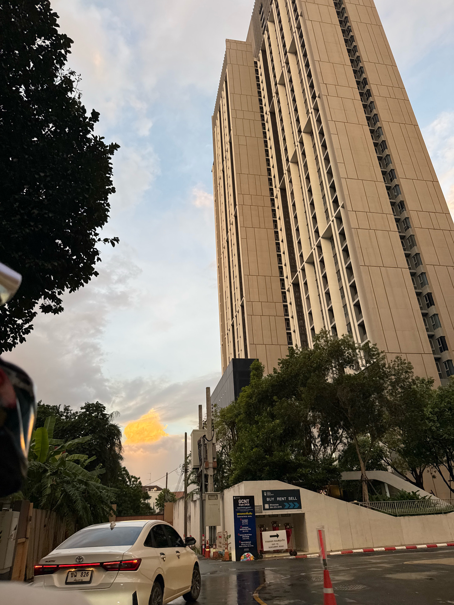 The image shows a low-angle view of a tall, modern residential building in Bangkok, Thailand. The building is light beige and features numerous repeating windows and balconies. In the foreground, a white sedan is parked on a wet street, suggesting recent rain. To the left, a portion of a dark green tree canopy is visible, and to the right, there are more trees and a white concrete structure with an entrance labeled "BUY RENT SELL." The sky is cloudy with patches of orange and yellow light, indicating either sunrise or sunset. The overall scene depicts an urban environment with residential architecture and signs of recent weather.