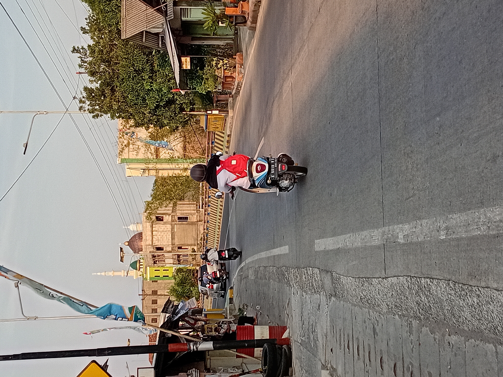 The image depicts a street scene in Bae, Indonesia, with a focus on a person riding a scooter. The scooter rider is wearing a black helmet and a white shirt with a red backpack. The scooter itself is blue and white, with a visible license plate displaying "K 6019 VT". The scooter is traveling on a paved road, marked with white lines.
In the background, several buildings are visible, including what appears to be a mosque with green and brown domes and a tall minaret. Construction is ongoing on some of the buildings, with exposed brickwork. Trees and vegetation line the side of the road, and power lines are strung overhead.
To the left of the road, there are some small structures, possibly shops or stalls, with people sitting near them. A yellow traffic sign is also visible on a pole. The lighting suggests it is late afternoon, with the sun casting long shadows. The overall impression is of a typical street in a moderately developed urban or semi-urban area.