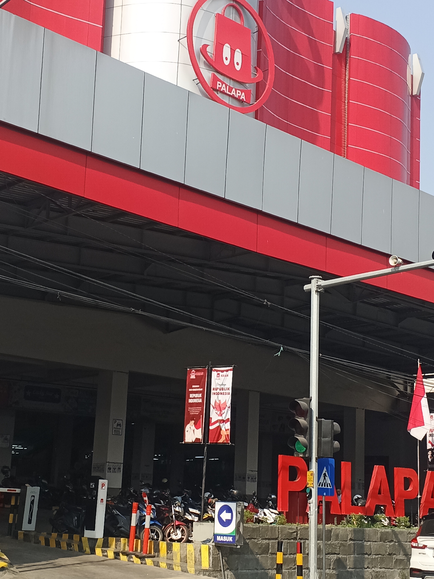 This image captures the exterior of a Palapa branded building in Kemangsen, Indonesia. The building features prominent red and grey architectural elements. Above the entrance level, a large, circular red logo with a shopping bag icon and the word "PALAPA" is visible. Below this, a wide grey band runs horizontally across the facade, separated from the lower red section by a thin red line.
The ground level appears to be a parking area, with several motorcycles and a few cars visible. A traffic light is positioned on a pole to the right of the frame, with its green light illuminated. A large, red, three-dimensional sign spelling out "PALAPA" is placed in front of the building. Two red and white banners hang near the traffic light, one displaying "DARING REPUBLIK INDONESIA." A pedestrian crossing sign is also visible near the large red lettering. The sky is clear and blue, suggesting a sunny day. The overall impression is that of a commercial establishment with a visible branding presence.
