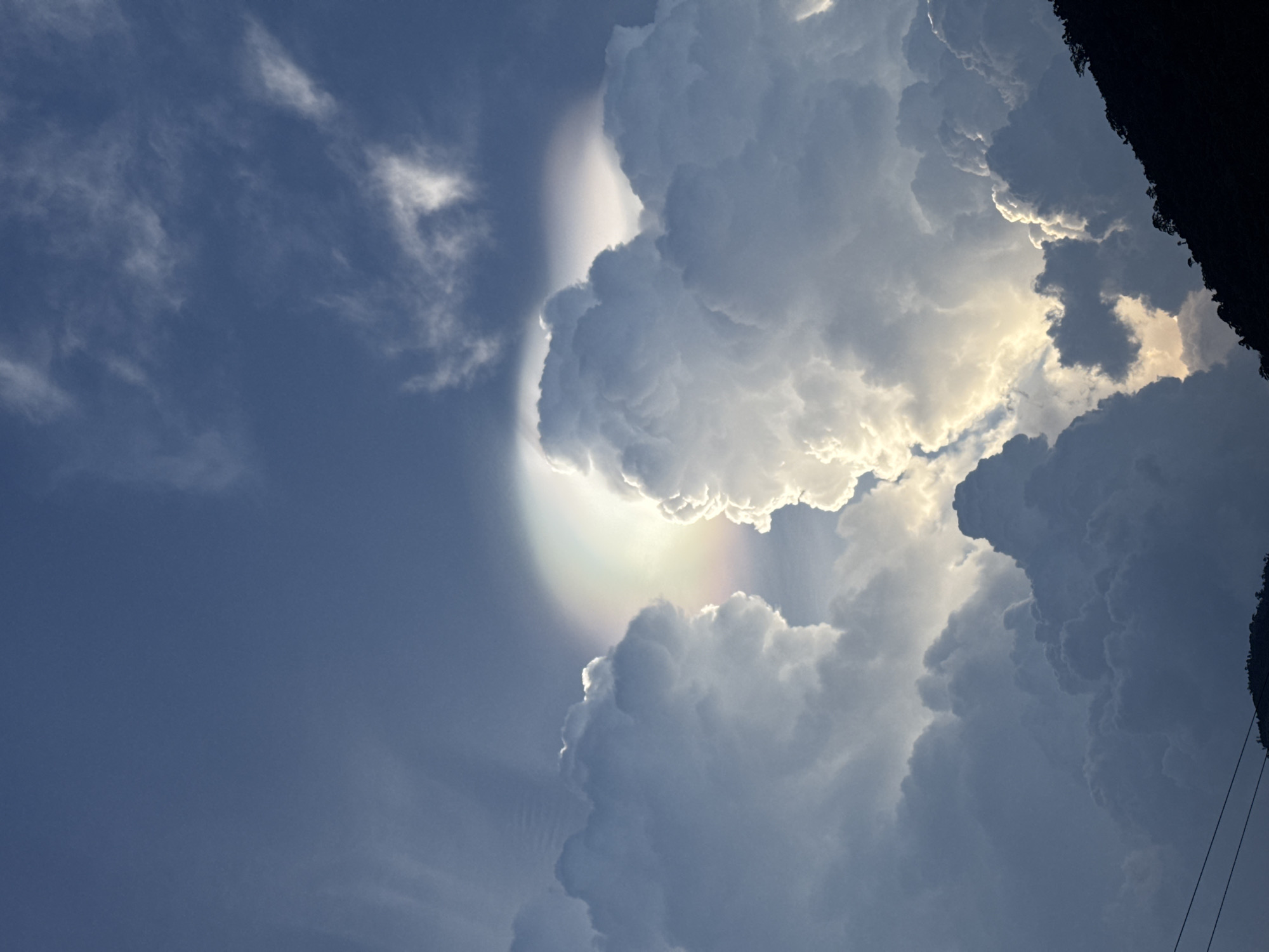 The image depicts a view of the sky in Tay Ninh Hoa, Vietnam. Large, billowing cumulonimbus clouds dominate the right side of the frame, some brightly illuminated by the sun from behind. A faint rainbow-like iridescence is visible in the clouds near the center. To the left of the clouds, the sky is a clear blue with thin, wispy clouds scattered about. Part of a dark structure, possibly a building or trees, is visible at the top right corner of the image. Electrical wires are faintly visible running down the right side. The viewpoint is looking upward.