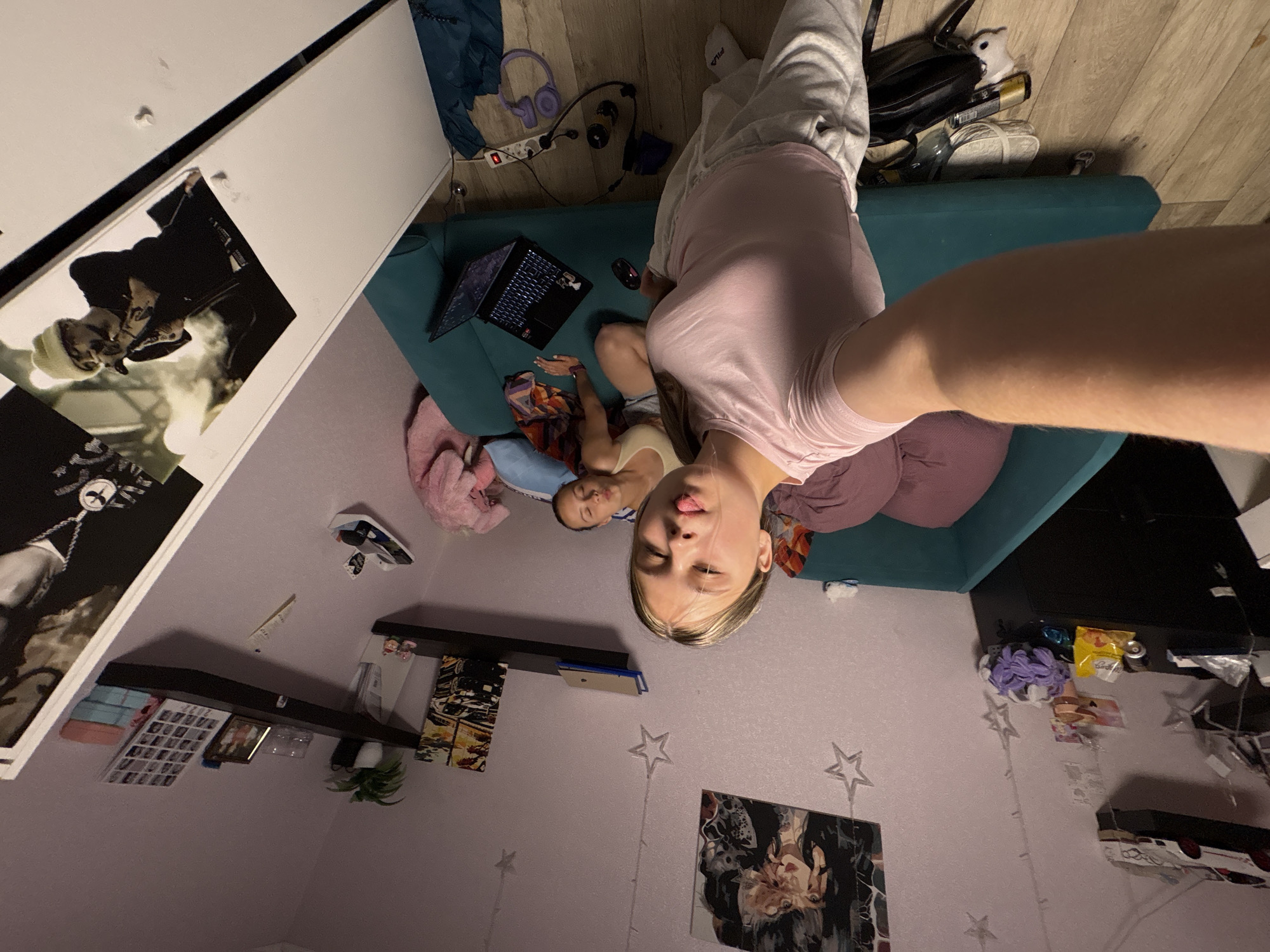 In a residential room in Прикамский, Russia, a young female with light hair takes a selfie from a high angle, extending her right arm. She wears a pink t-shirt and grey sweatpants, with a "FILA" logo visible on her left sock, and her tongue is slightly out. Behind her, a second young female with dark hair rests on her side on a teal sofa, partially covered by a patterned textile. A pink plush toy is visible near the second female on the sofa.
The room features light purple walls adorned with star-shaped string lights and various artworks, including a stylized portrait of a woman and a depiction of a car. Floating shelves display model cars, a potted plant, framed photos, a photo grid possibly dated "2022," and stacked towels. A black dresser on the left holds a yellow snack bag and a purple ball of yarn. On the wooden floor near the sofa, a black laptop with an illuminated keyboard, a computer mouse, and purple headphones are scattered. A white power strip and a dark blue piece of fabric are also visible on the floor in the foreground. A large print of a man wearing a beanie and a necklace, holding a microphone, is positioned vertically on the far right.