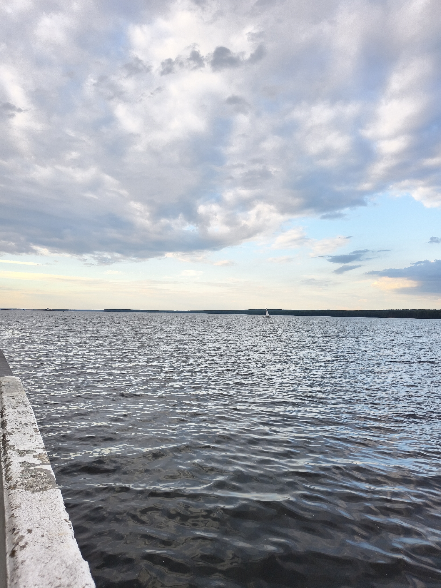 The image shows a wide expanse of water, likely a reservoir or large lake, with a distant treeline on the horizon. The sky above is filled with a mix of white and gray clouds, with some patches of blue visible. In the middle distance, a small white sailboat with a single sail is visible. The foreground features a concrete barrier or walkway with a rough, weathered texture. The water is rippled, indicating a light breeze. This scene is consistent with views of the Kama River reservoir near Chaikovsky, Russia, known for its large bodies of water and recreational activities like sailing. No people are visible in the image.