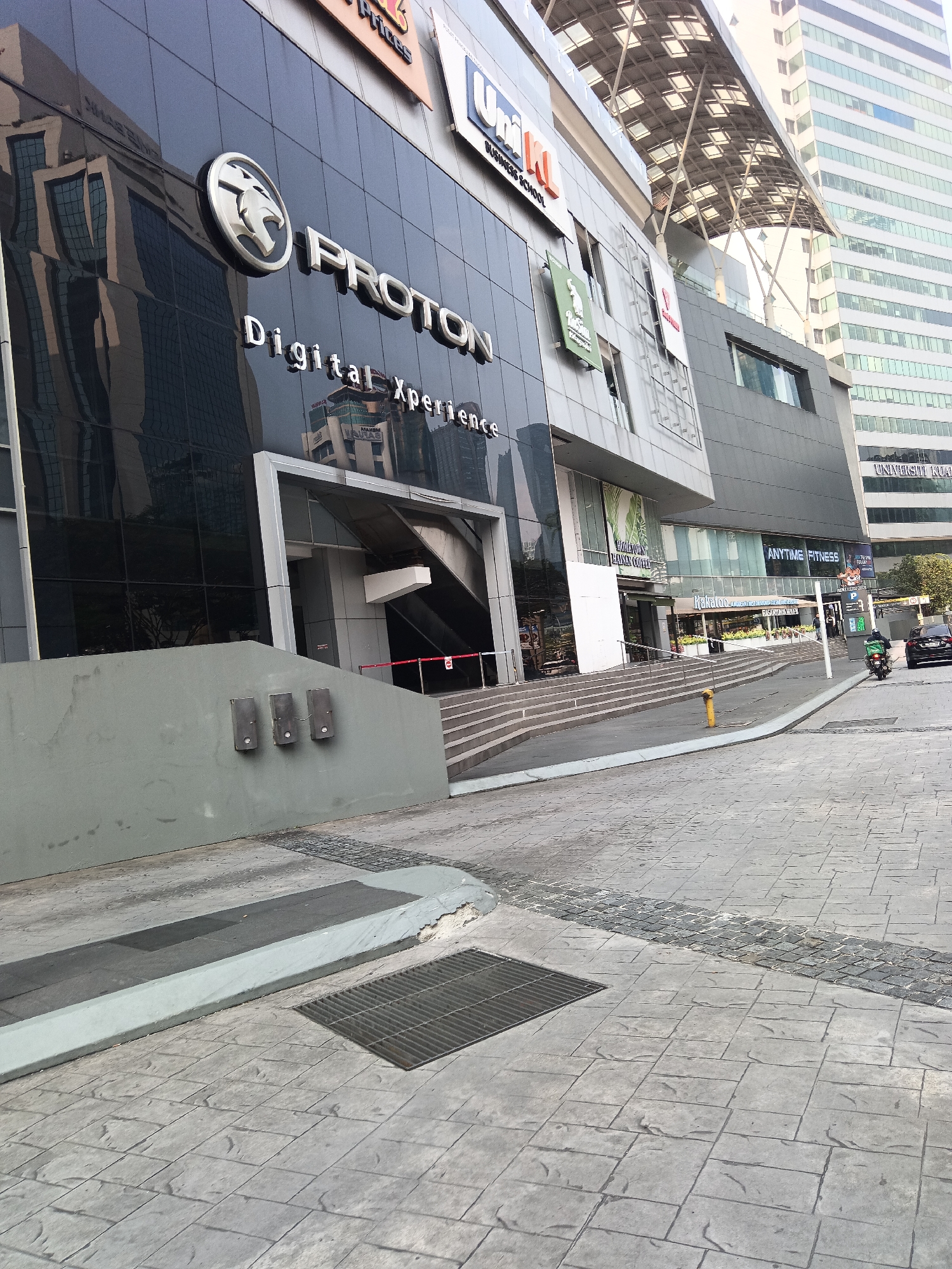 The image shows the Proton "Digital Xperience" showroom located in Kuala Lumpur, Malaysia. The exterior of the building features a modern architectural design with a black glass facade. The Proton logo and name are prominently displayed on the building. Reflections of surrounding buildings can be seen in the glass. In front of the showroom, there is a paved area with patterned concrete, a ramp leading up to the entrance, and some outdoor electrical boxes mounted on a grey wall. Further to the right, the entrance to other businesses such as "Anytime Fitness" and "Kukubotan" is visible. There is a street with a few vehicles, including a motorcycle with a delivery rider and a car, suggesting it's a commercial or mixed-use area of the city. The overall scene captures a street-level view of a commercial establishment in an urban environment.