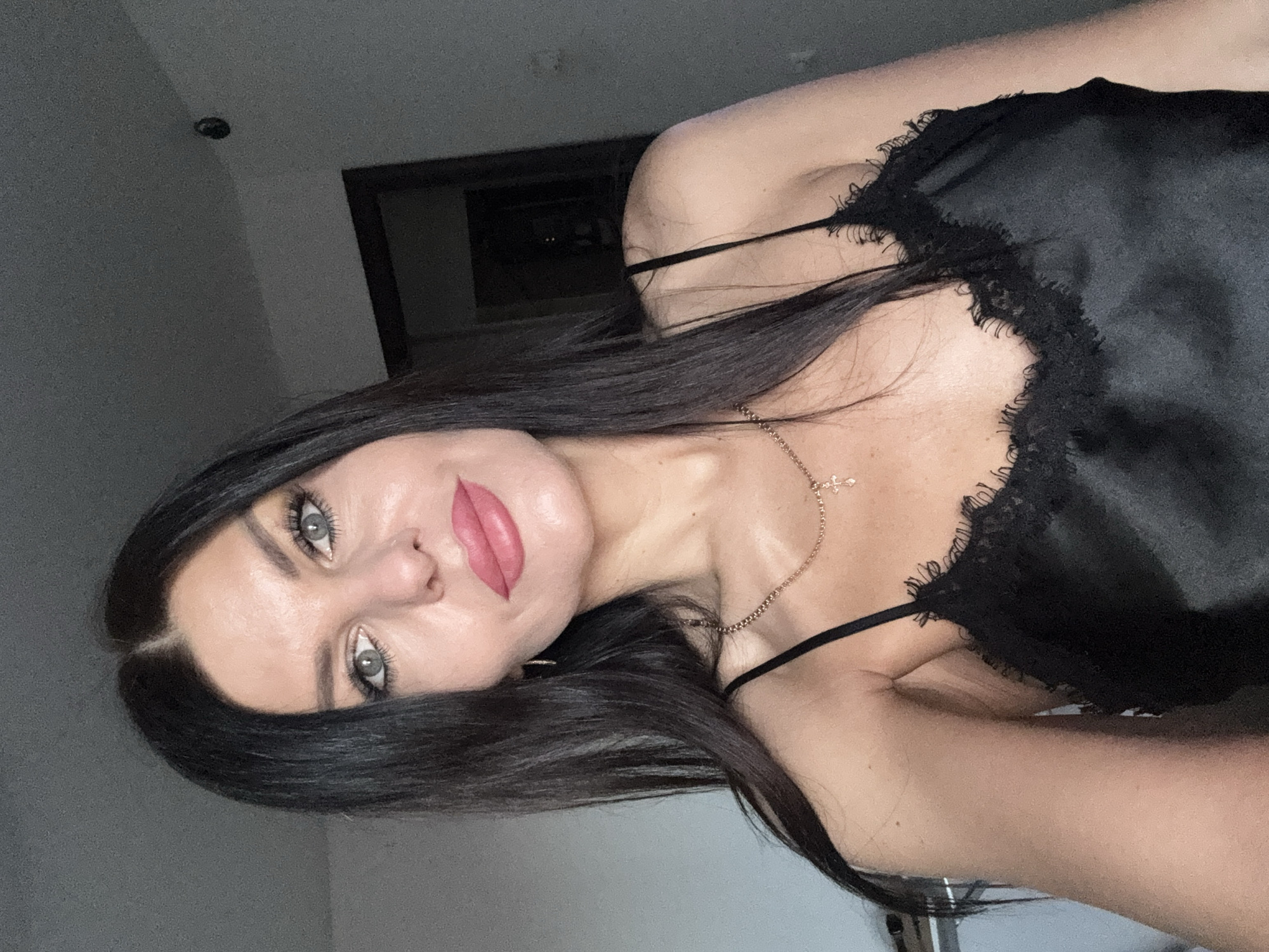 The image is a vertical portrait of a woman indoors. She has long, dark hair, prominent eyebrows, and striking green eyes. Her lips are painted with a vibrant pink lipstick. She appears to be in her 20s or 30s.
The woman is wearing a black, satin or silk top with delicate lace trim along the neckline and thin spaghetti straps. A delicate gold chain necklace with a small cross pendant hangs around her neck, resting on her collarbone.
She is looking directly into the camera with a neutral to slightly contemplative expression, suggesting a posed photograph or selfie. The lighting is soft, creating highlights on her skin, particularly on her chest and shoulders.
The background is simple and somewhat indistinct, featuring a light grey wall or ceiling on the left, and a darker area on the right that appears to be a doorway or an opening into another room. A small, dark light fixture is visible on the ceiling in the upper left corner of the frame. There is no visible text in the image.
