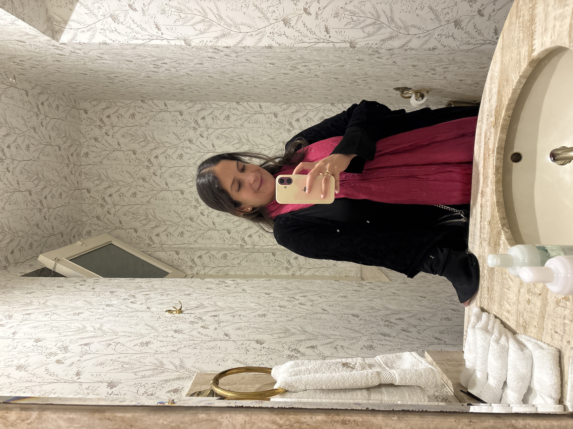 An individual stands in front of a mirror in a bathroom, taking a selfie. They are wearing a black cardigan over a black top and a bright pink scarf. The bathroom features light-colored, patterned wallpaper with a delicate botanical design. A gold towel ring with folded white towels hangs on the left side of the frame. The vanity is made of marble, and a sink is visible in the foreground, with several bottles of toiletries on the counter. The lighting suggests an indoor environment. The location is specified as Lima, Peru.